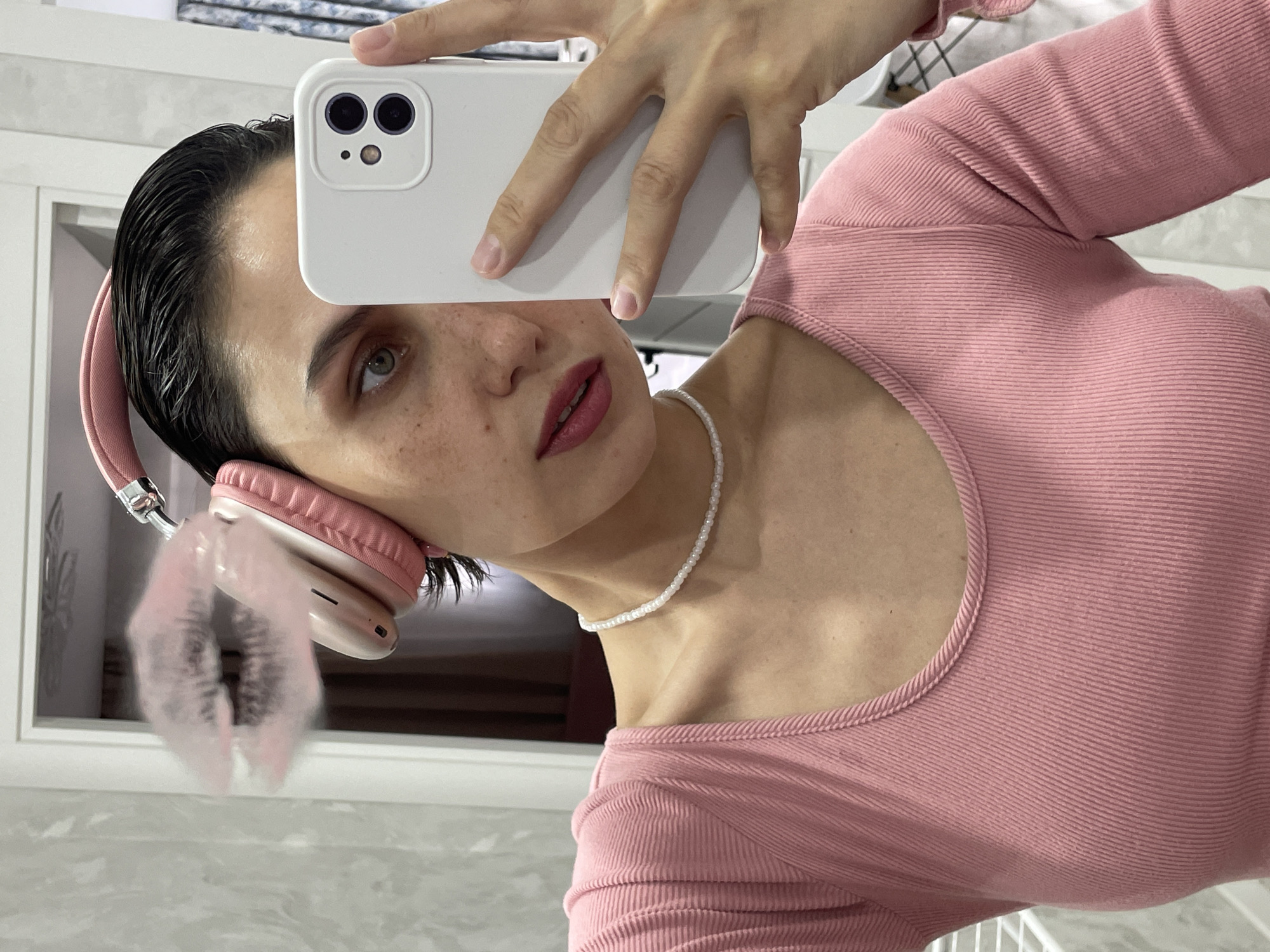 A person is taking a selfie. They are holding a white iPhone with a dual-lens camera. The person has dark hair slicked back, pink headphones, and freckles. They are wearing a pink ribbed top and a white beaded necklace. The background appears to be a room with white walls. There is no discernible location in Izhevsk, Russia visible in the image.