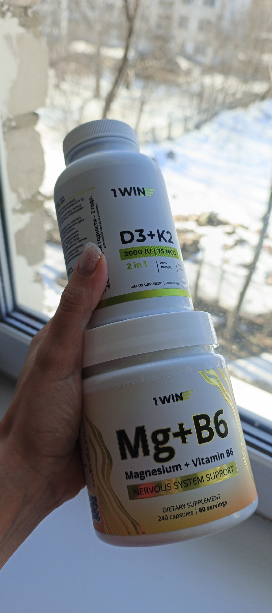 The image shows two bottles of vitamins in a person's hand. The top bottle is white and labeled "1WIN D3+K2" with the text "2000 IU / 75 MCG 2 in 1 Bone strength DIETARY SUPPLEMENT 180 capsules". The bottom bottle is white and labeled "1WIN Mg+B6 Magnesium + Vitamin B6 NERVOUS SYSTEM SUPPORT DIETARY SUPPLEMENT 240 capsules | 60 servings". The person's hand is holding the bottles in front of a window. Through the window, bare trees and a building are visible. According to text on the bottle, the image was taken in Chaikovsky, Russia.