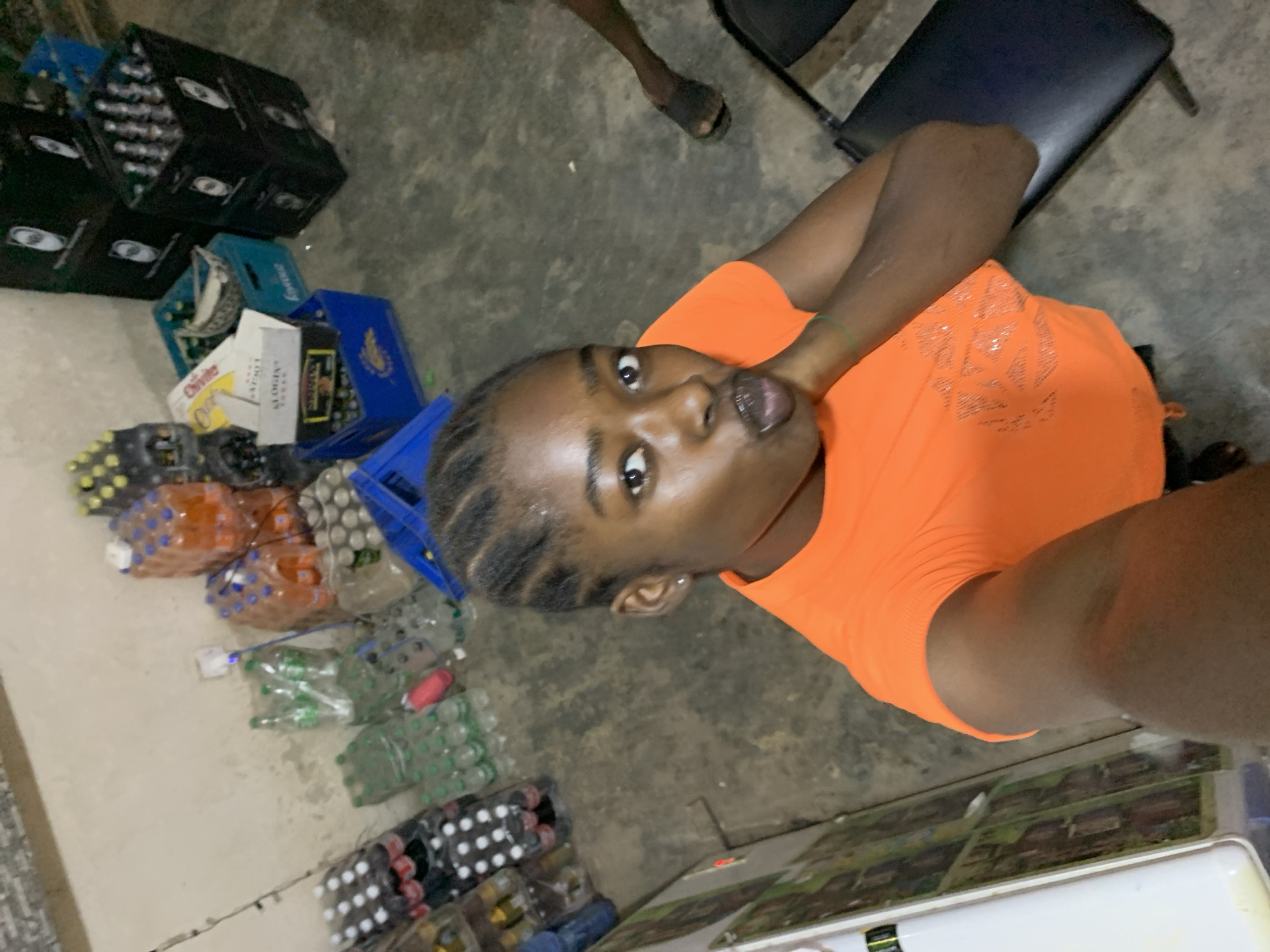 The image appears to be taken from a high-angle perspective in what looks like a small storage or warehouse area, possibly a shop or business based on the stacked drinks. The setting is likely in the city of New Stadium, Nigeria.
A young adult with an orange top is in the foreground, facing the camera. The floor is a light grey concrete. In the background are numerous stacked crates and packaged drinks, including bottled water, soft drinks, and possibly alcoholic beverages. A partial view of a black chair and other individuals' legs are present, indicating a gathering of people.