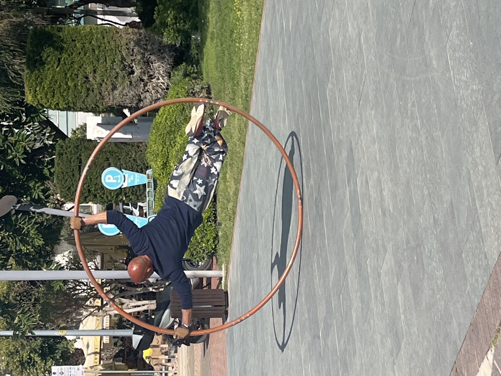 A man is performing inside a large metal hoop on a paved area in Alanya, Turkey. He is wearing a long-sleeved dark blue shirt, patterned pants, and brown gloves. The man is positioned inside the hoop, using his arm to help maintain his balance. The ground is paved with gray tiles. In the background, there are trees, bushes, and street signs, including one with a "P" inside a blue circle. A shadow of the hoop and the man is visible on the pavement.