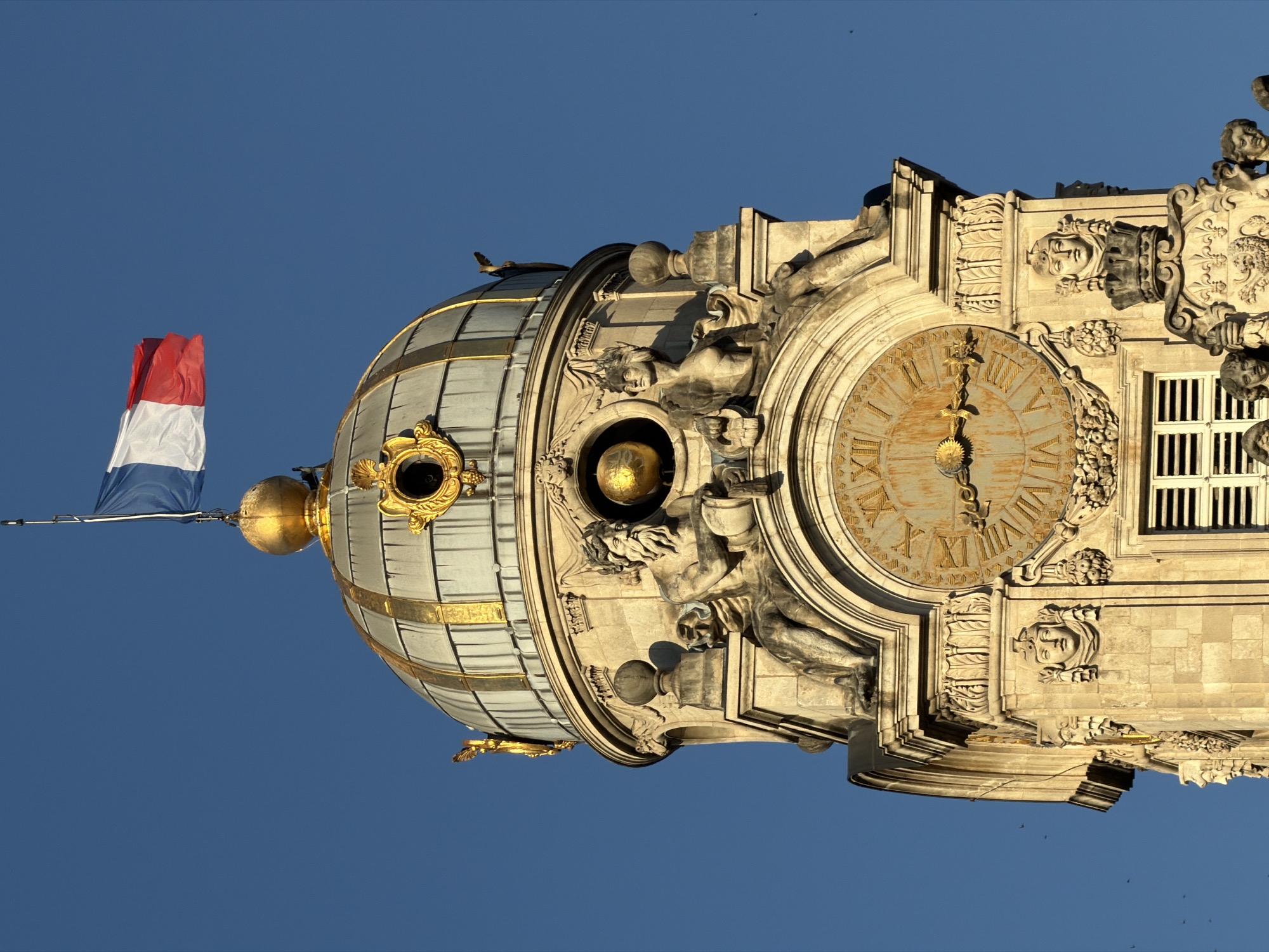 The image is a close-up, eye-level shot of the top of a historic clock tower in Lyon, France. The tower is made of stone and adorned with intricate sculptures and gold accents. At the very top, a French flag waves against a clear blue sky. Below the flag is a golden orb and the dome of the tower, which is decorated with gold trim. A large clock face with Roman numerals is prominently featured on the tower, with golden hands indicating the time. Statues of male and female figures, along with detailed carvings of faces and ornate architectural elements, surround the clock. The overall impression is one of grandeur and historical significance.