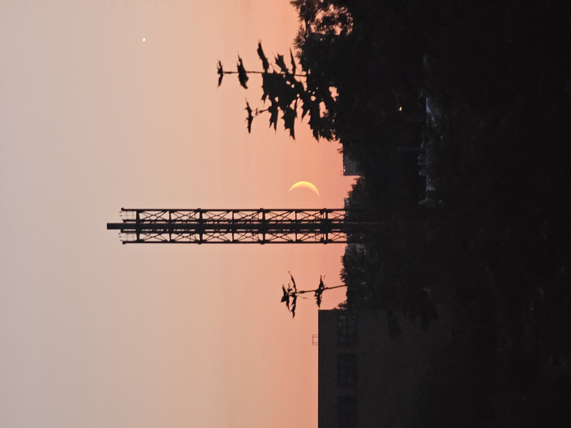 This is a vertically oriented, medium shot of a telecommunications tower silhouetted against a pale orange sunset sky, taken in Khimki, Russia. The tower, located in the mid-right of the frame, is tall and lattice-like. To the left of the tower, a crescent moon is visible. In the foreground and mid-ground, the silhouettes of trees and buildings obscure much of the lower half of the image. A large, dark rectangular building is visible to the left. The sky is clear and shows a gradient from a lighter shade at the top to a deeper orange near the horizon. A small, bright point, likely a star or planet, is visible in the upper right quadrant of the sky. The overall mood is one of twilight.