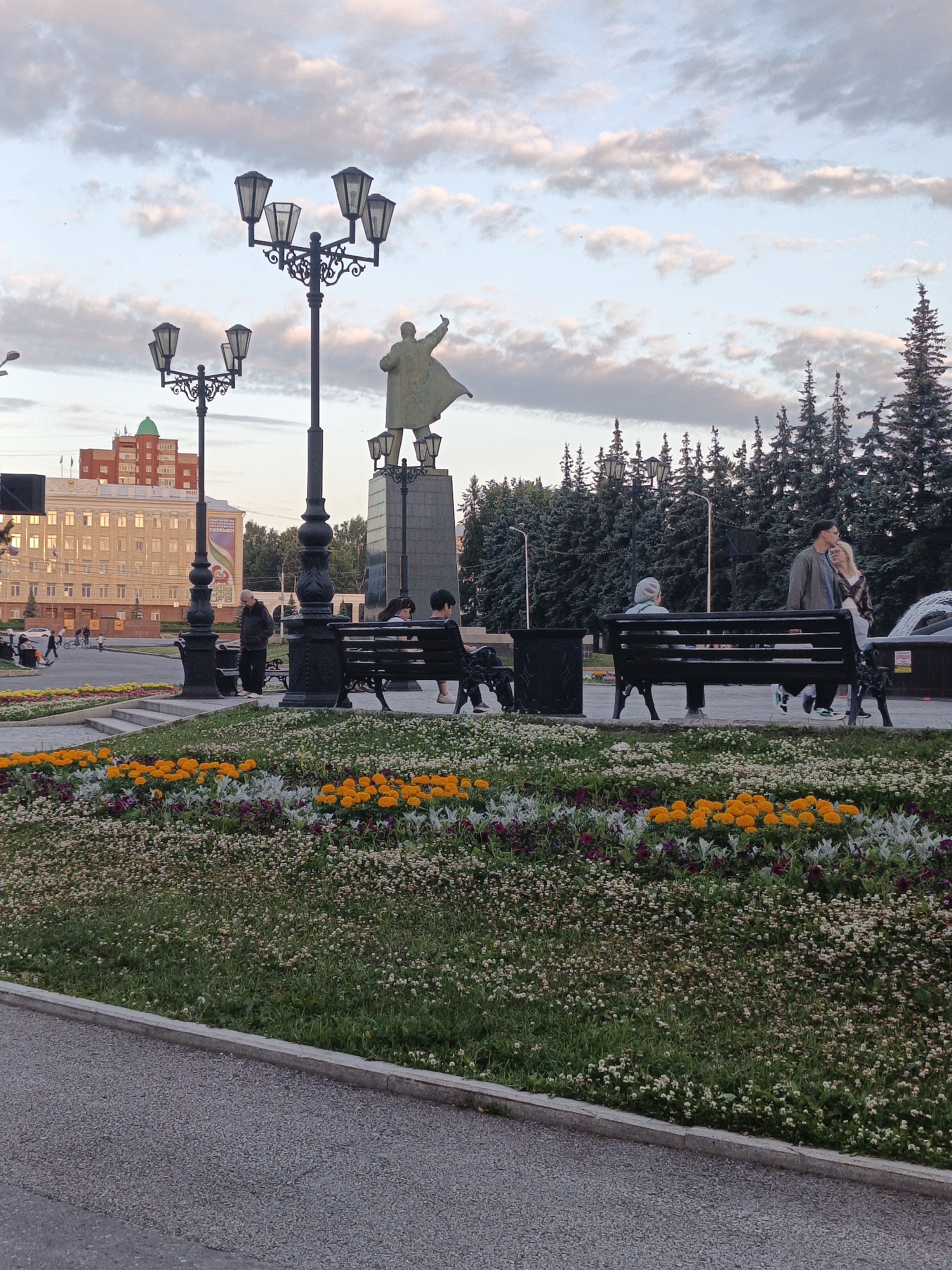 The image depicts a square in Ufa, Russia, featuring a statue of Lenin in the background, pointing towards the sky. The foreground is adorned with a vibrant flower bed containing yellow, purple, and white blossoms. Several people are seated on benches, enjoying the evening atmosphere. A couple is captured in a tender moment, while others are engaged in conversations or simply relaxing. The presence of lampposts and a distant building suggests an urban setting. The sky is a canvas of pastel clouds, indicating a serene evening. The overall scene portrays a peaceful communal space, likely a popular gathering spot in the city.