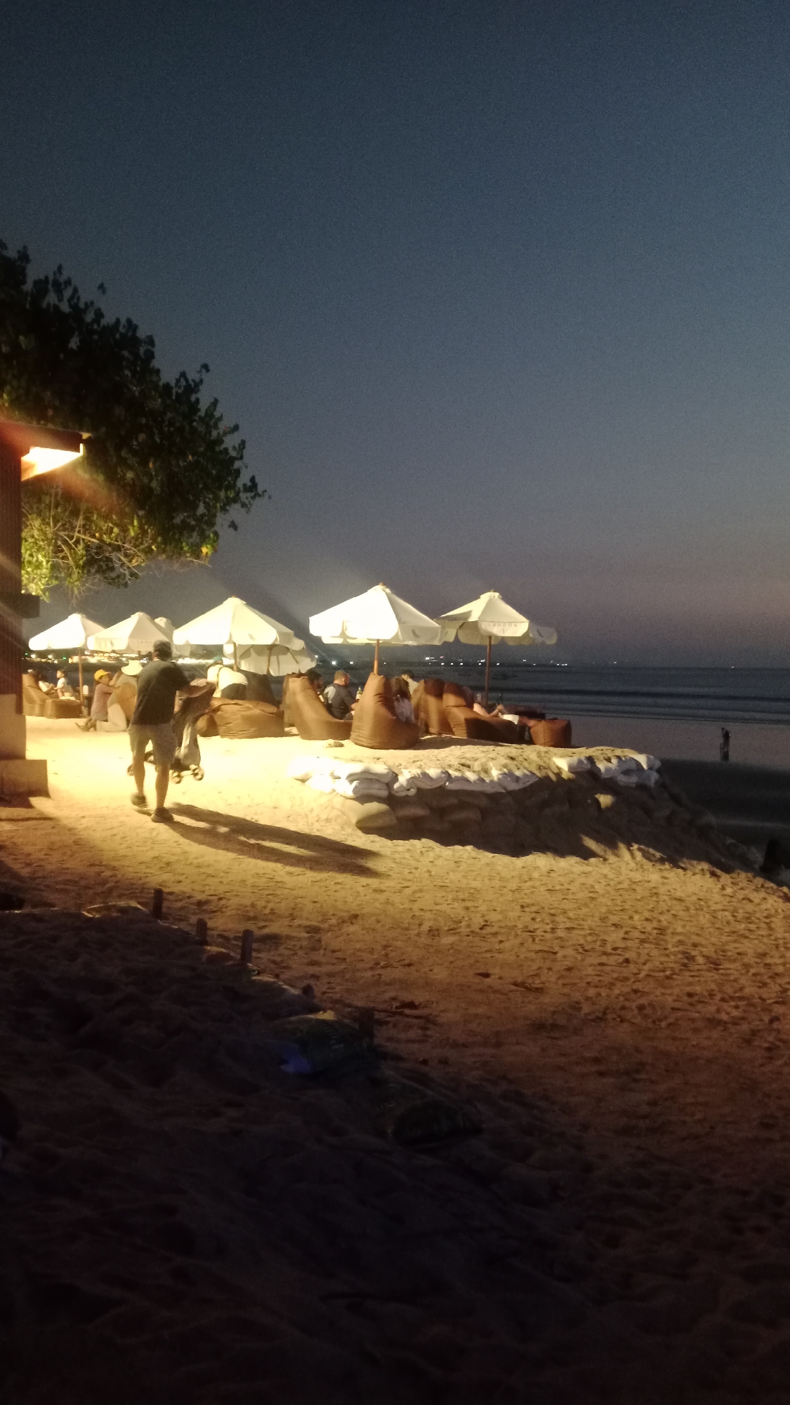 The image depicts a beach scene at dusk in Kuta, Indonesia. The sand is illuminated by artificial lighting from the left, casting long shadows. Several white umbrellas are set up, providing shade for beanbag chairs where people are seated, relaxing. A man walks away from the camera on the left side of the image, appearing to be carrying something. The ocean is visible in the background, with distant lights on the horizon. The sky is a deep twilight blue, transitioning to a lighter shade near the horizon. The overall atmosphere is relaxed and serene, typical of a beachside lounge area in Kuta.