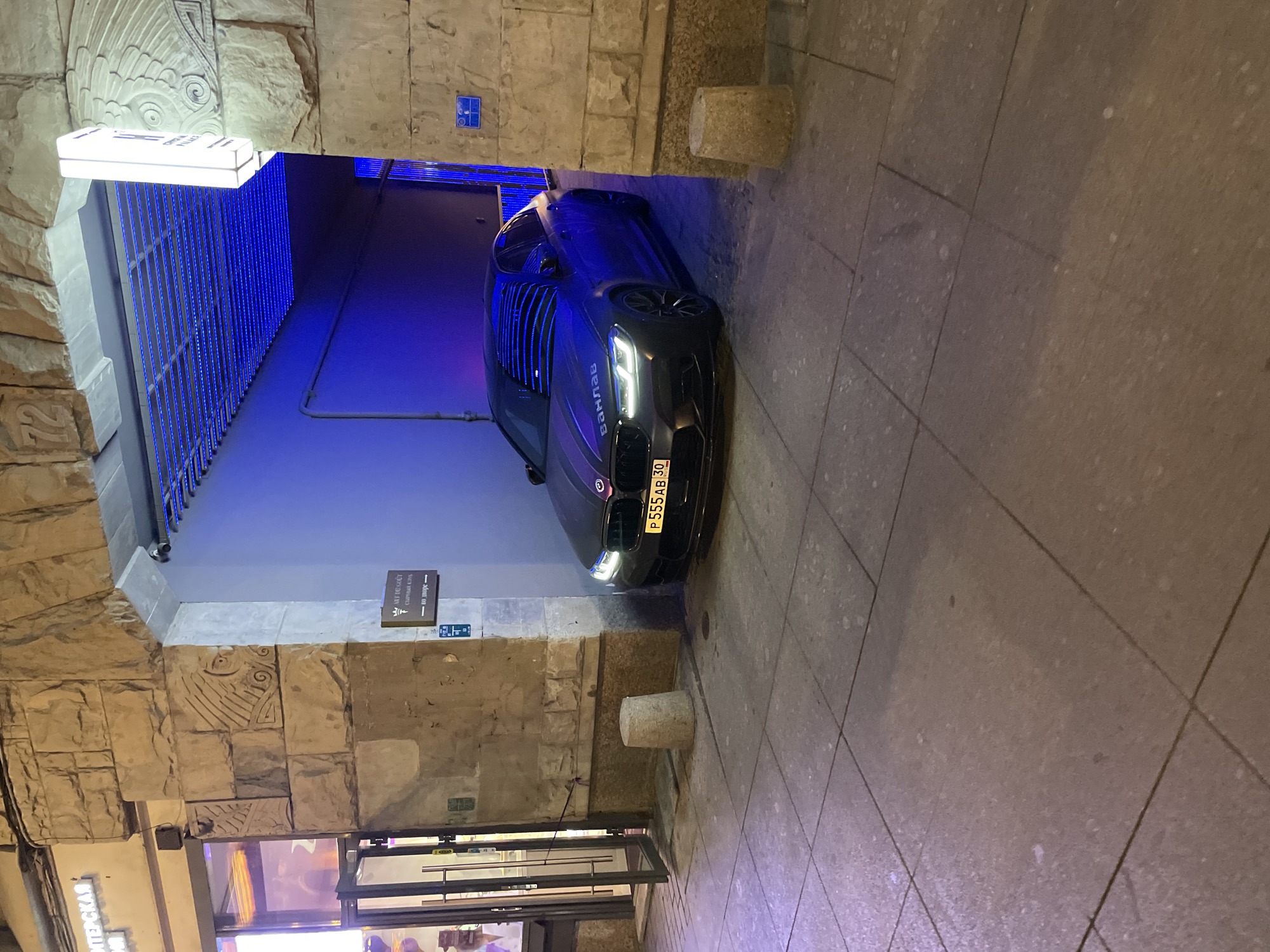The image depicts a nighttime urban scene in Saint Petersburg, Russia, focusing on a dark-colored BMW M5 sedan (F90 generation) partially emerging from or entering a stone archway. The vehicle, dark gray or matte black with a purple-edged black stripe on the hood and the Cyrillic text "ВАНЛАВ" (VANLAV) emblazoned on it, displays Russian license plate P555AB 30. Its headlights are illuminated.
The archway, constructed of roughly cut stone blocks, features decorative carvings: an owl-like figure on the left pillar and a winged ornament on the right pillar. A prominent "72" is carved into the stone above the archway's apex. The interior of the archway is dramatically lit with an array of blue LED strip lights mounted on a grid ceiling structure, casting a strong blue hue on the side walls.
To the immediate left of the archway, a commercial establishment with a modern glass entrance and double doors is visible. Above its entrance, a glowing sign displays partial Cyrillic text, appearing to be "ИНТЕРСКАЯ" (INTERSKAYA). On the right stone pillar of the archway, a brown rectangular sign points left with an arrow and reads "ART DU GOÛT СЫРНЫЙ КЛУБ" (Cheese Club Art Du Goût) above "← ВО ДВОРЕ →" (To the courtyard). Below this, a smaller blue plaque is affixed. Further right on the same pillar, a vertical white sign with internal illumination reads "CRYSTAL PALACE" in English, with additional Cyrillic text below. Another small blue plaque is visible beneath the "CRYSTAL PALACE" sign.
The foreground consists of a sidewalk paved with large, rectangular, light gray stones. Two cylindrical concrete bollards are present on the sidewalk: one on the left near the shop entrance and another partially visible on the right next to the car. The lighting is artificial, comprising the blue LEDs within the archway, the glowing shop sign, and general ambient street lighting. No individuals are visible in the scene.
Based on the combination of signs for "ART DU GOÛT СЫРНЫЙ КЛУБ" and "CRYSTAL PALACE" located at a distinctive archway with carved stone elements, the location is identified as **Karavannaya Ulitsa, 7, Saint Petersburg, Russia**. The visible "72" above the archway may refer to an internal building section number or an older numbering system.