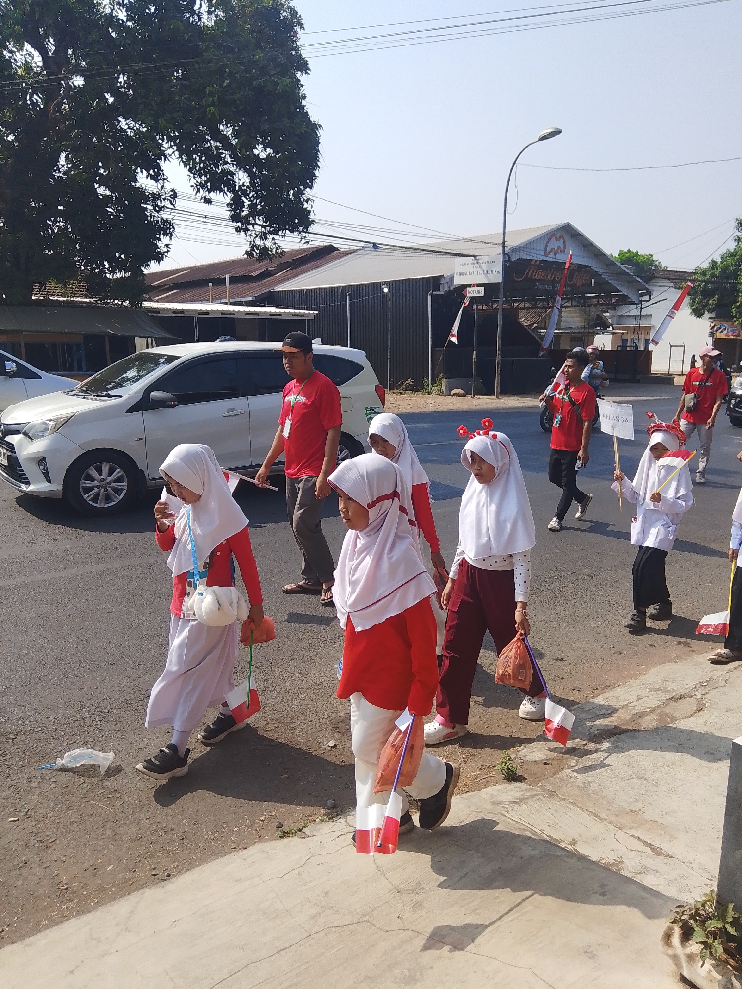 In Kalinyamatan, Indonesia, on a sunny day, a group of schoolchildren in red and white uniforms are participating in a parade. Some of the children are carrying small Indonesian flags. A man in a red shirt is walking behind them, possibly supervising. A white car is parked on the side of the road, and buildings are visible in the background, including one with a sign that reads "Maestro Coffee". The scene appears to be taking place on a street with a sidewalk.