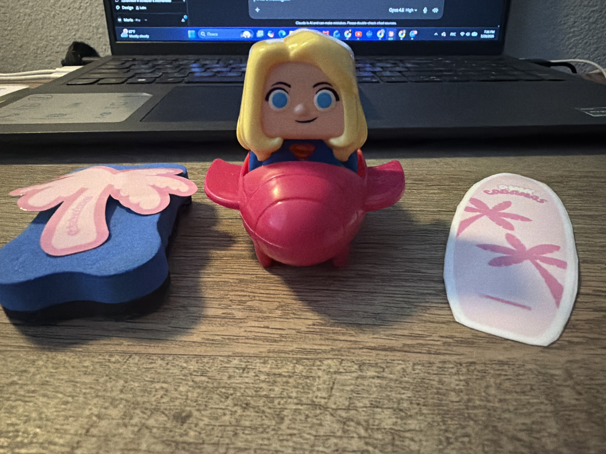 A Supergirl figurine in a pink airplane toy is positioned in the center of the frame, resting on a wooden desk. To the left of the figurine is a blue foam block with a pink flower sticker. To the right of the figurine is a pink and white oval-shaped sticker with a pink flower design. In the background, a laptop computer is visible. The scene is indoors, and no specific location in the city of Orlando, United States of America is identifiable.