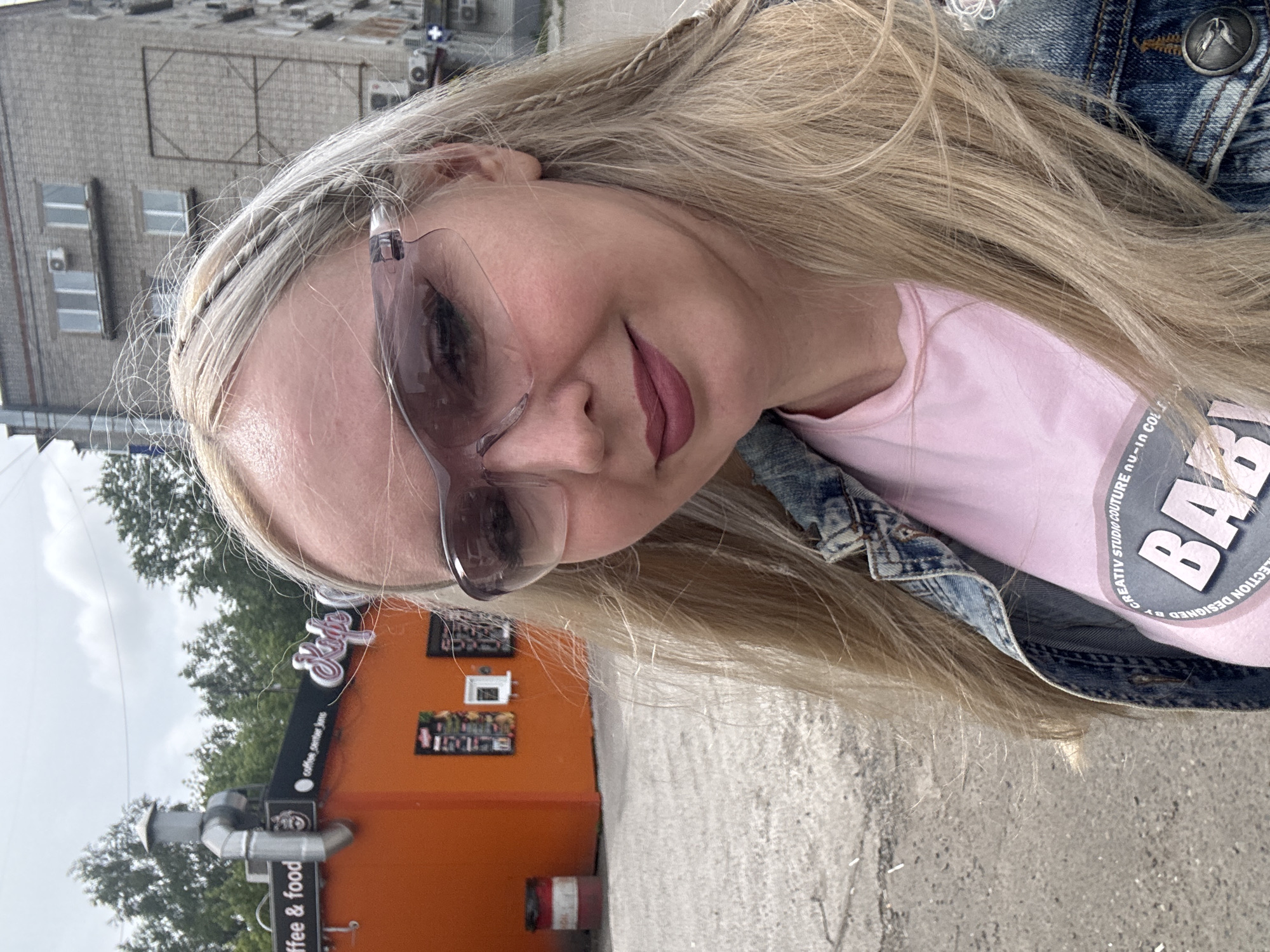 A woman with long blonde hair, braided in sections, wears a denim jacket over a pink t-shirt. She has on large, pink-tinted glasses and dark lipstick. She is outdoors in front of an orange building that appears to be a food stand or cafe, with a sign reading "coffee & food." To the right of the building is a brick apartment complex. The ground is paved and appears to be a parking lot or walkway. The sky is overcast. The image was taken in Komsomolsk-on-Amur, Russia.