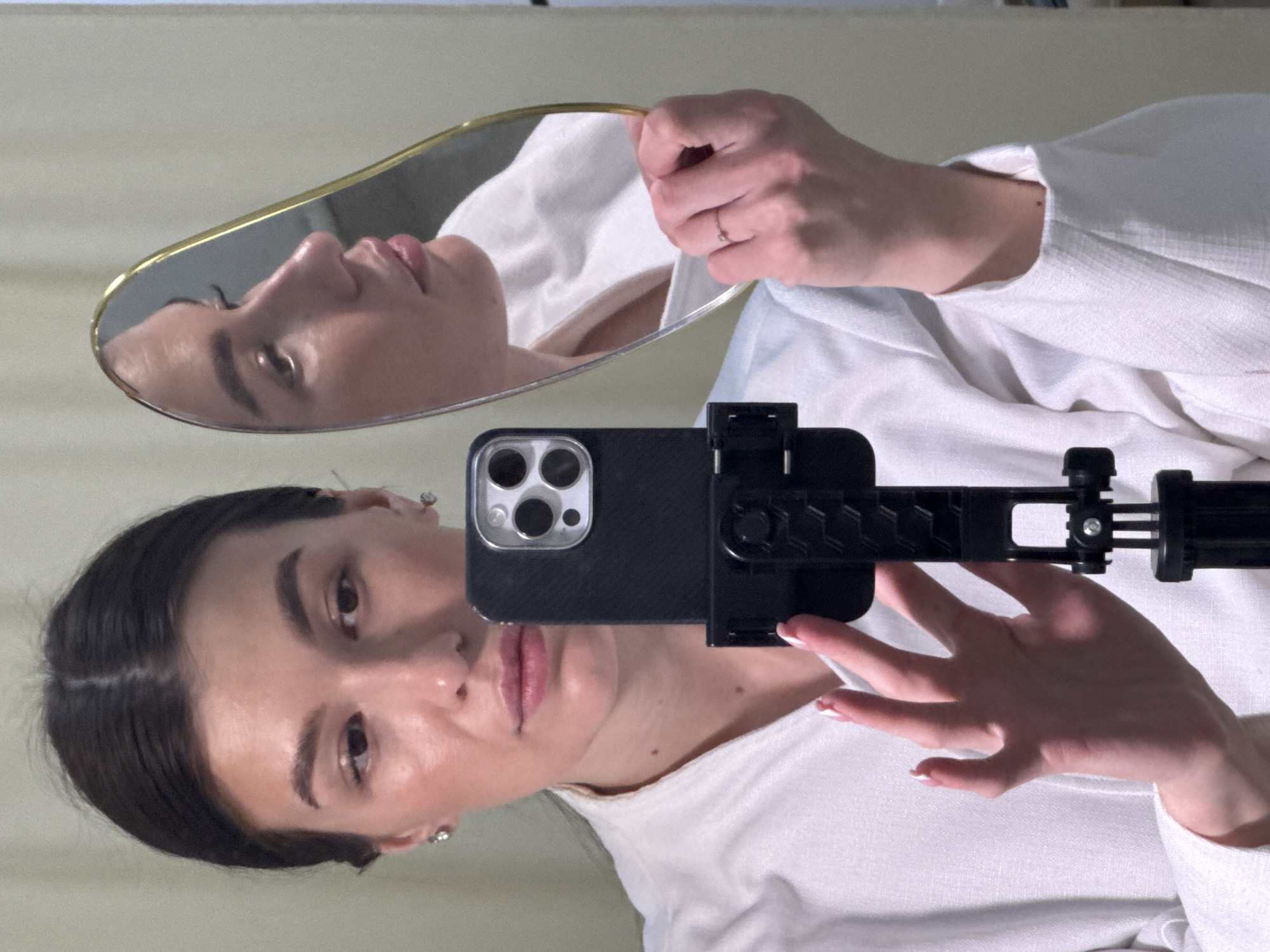 A self-portrait captured indoors shows a person, likely a young adult female, rotated 90 degrees clockwise within the frame, appearing to lie on her side. She is engaged in a complex self-documentation process, holding a smartphone mounted on a black tripod-style holder with her right hand, seemingly using it to take a photo or video. Simultaneously, her left hand holds a small, ornate handheld mirror with a golden rim, reflecting her profile.
The subject has dark hair with a center part, pulled back, revealing small stud earrings. She possesses prominent eyebrows and a neutral expression with slightly parted, full lips. Her complexion appears clear, with subtle highlights on her cheekbones and nose. The reflection in the mirror shows her profile, emphasizing her nose and lips. A small ring is visible on a finger of the hand holding the mirror. Her nails feature a neat French manicure.
She is wearing a white, long-sleeved garment, possibly a shirt or top, made of a textured fabric. The smartphone is a modern iPhone, identifiable by its triple-lens camera array and flash module, housed in a black protective case. The background is an indistinct, plain light-colored surface, suggesting an indoor setting. The lighting is soft and even, without harsh shadows, indicative of artificial or diffused natural light. The overall mood is contemplative and artistic, focusing on self-perception and reflection. The location context for this media file is Chaikovsky, Russia. There is no visible text in the image.
