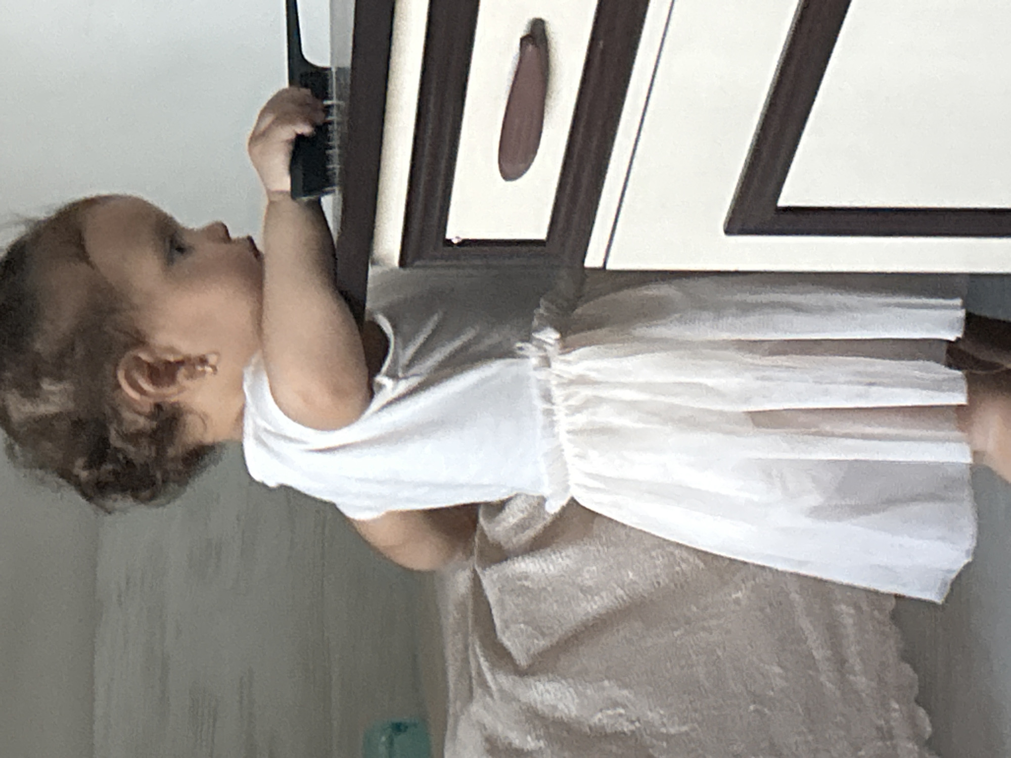 The image shows a young child, likely a baby or toddler, leaning towards a piece of furniture. The child has curly brown hair and is wearing a white top with ruffled sleeves and a white tutu-like skirt. The child's left hand is reaching out towards the furniture, holding a dark object that appears to be a comb or a similar grooming tool. The furniture is light-colored with dark brown trim and a small, oval-shaped handle in the center of one of the panels. The setting appears to be indoors, possibly a bedroom or living area, with neutral-colored walls and a floor with a textured pattern. The lighting suggests it is daytime. There is no visible text in the image. The child's expression is focused on the object they are interacting with.