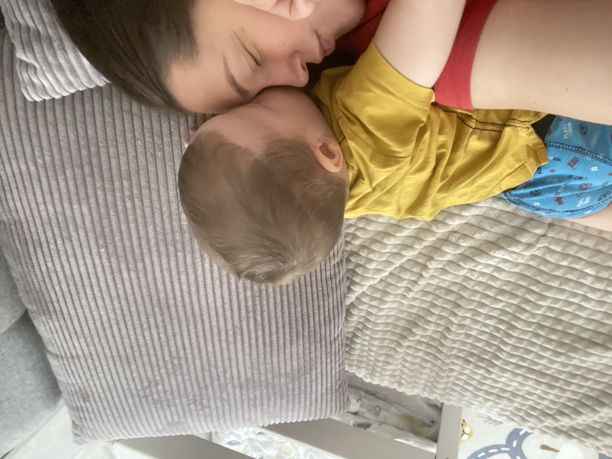 An aerial, close-up shot captures a person and a baby lying together on what appears to be a bed, possibly in a residential setting. The adult has dark hair and is wearing a red shirt. The baby has light-colored hair and is wearing a yellow shirt with blue underpants patterned with white images. They are lying close together, and their faces are near each other. They are resting on a white textured blanket with what appears to be a gray corduroy pillow to the left. The lower portion of the frame contains a white-colored bedframe and a portion of a baby play mat patterned with streets and toy cars. The image does not contain information or cues to identify the location of the city of Sochi, Russia.