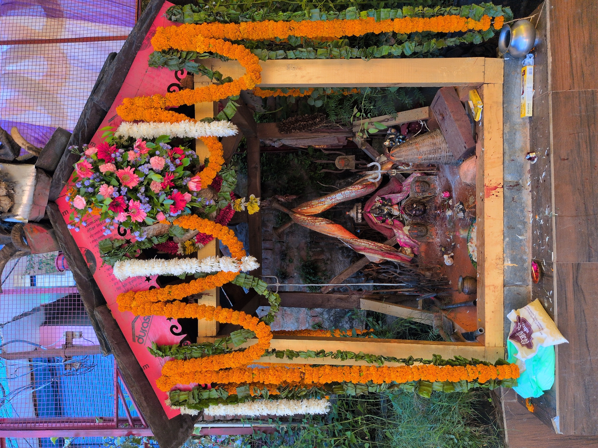 A small, open-air shrine dedicated to a deity, likely Kali, is depicted outdoors in Kullu, India. The shrine is adorned with vibrant orange marigold garlands and white flower garlands, as well as a lush bouquet of pink and purple flowers at the apex. The central figure within the shrine appears to be a dark idol with pronounced features, possibly adorned with fabric and jewelry. Several symbolic items are present, including a trident, a basket-like structure, and a coconut. The ground around the shrine is littered with debris, including what appears to be rice, incense sticks, and small offerings. In the foreground, a teal plastic bag and a packet of rice are placed on a wooden surface, alongside a small metal pot. The background features brickwork and foliage, suggesting an outdoor, possibly mountainous or forested, environment. The overall impression is one of a devotional space, possibly a roadside altar or a small temple setup, decorated for a religious ceremony or festival.