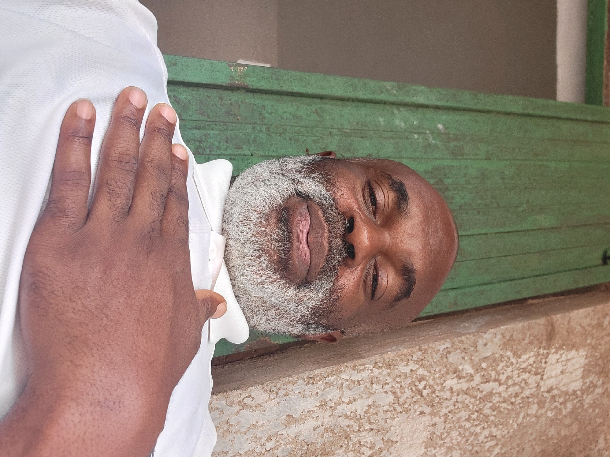 An intimate, portrait-oriented shot, taken in Maiburiji, Nigeria, depicts two individuals in what appears to be a casual setting. The image is composed with one person's hand prominently in the foreground, resting gently on the shoulder or chest of another. This person, whose hand is dark-skinned, is wearing a white collared shirt.
The main subject is a dark-skinned man who occupies the right side of the frame. He appears to be middle-aged or older, distinguished by a full, well-groomed grey beard and short, grey hair. He is also dressed in a white collared shirt, visible at his neck. The man looks directly or slightly upwards towards the camera with a calm, serene expression, featuring a subtle smile. His head rests against a rustic, distressed green surface, which appears to be made of wood with horizontal panels, possibly a door or a shutter. Below this green structure, a textured, light brown or beige wall or ground surface is visible, adding to the unadorned background.
The lighting in the image suggests daytime, but it is diffused, indicating either an indoor setting, an overcast day, or shade, as no harsh shadows are present. No discernible text is visible anywhere in the frame. The overall atmosphere conveys a sense of quiet presence and personal connection between the two individuals.