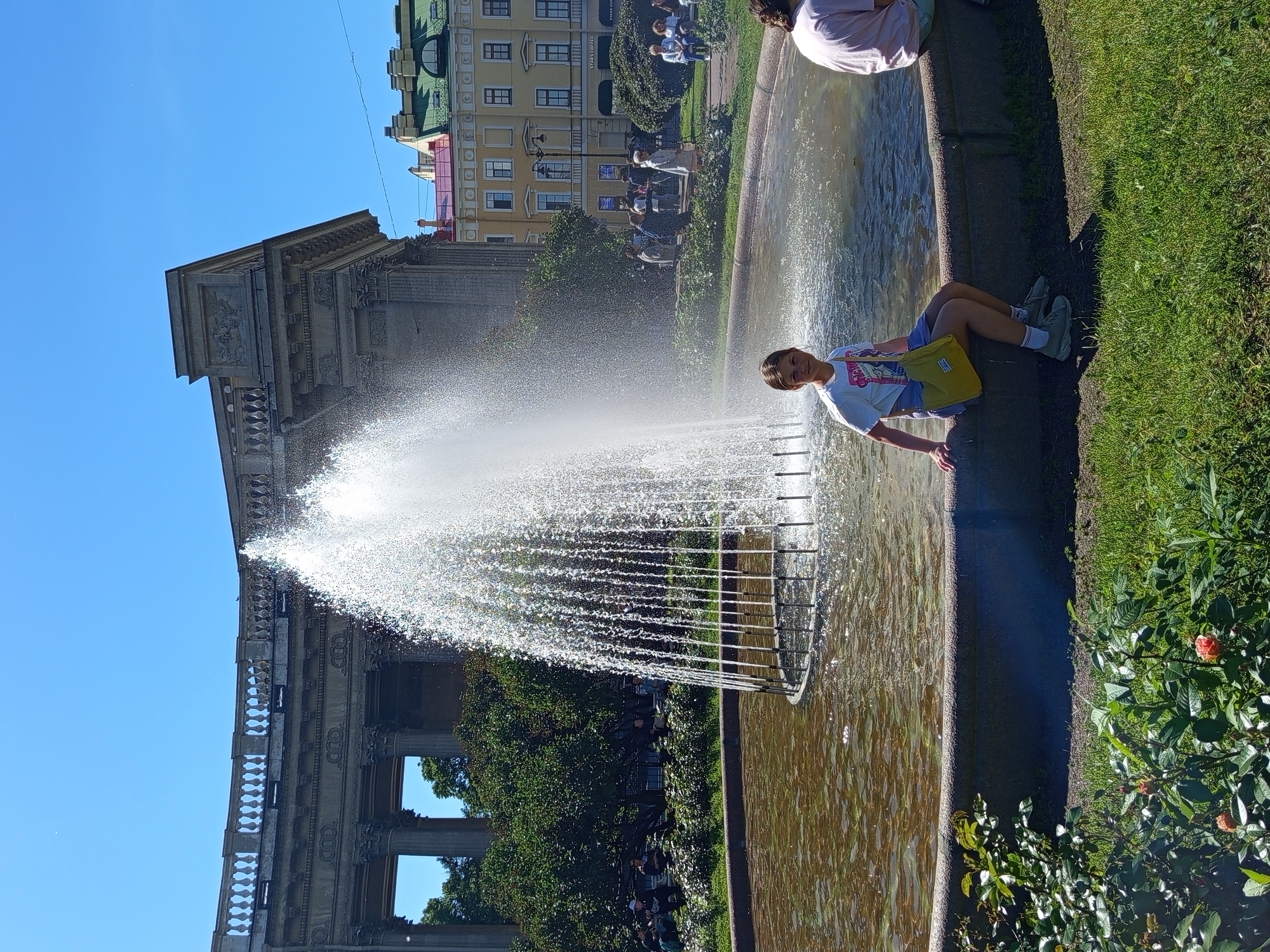 In Saint Petersburg, Russia, a young person sits by a fountain, holding a yellow bag. The fountain sprays water into the air, catching the sunlight. Behind the fountain, an ornate building with classical architecture stands under a clear blue sky. The building has columns and detailed carvings. Further in the background, there are more buildings with green roofs, suggesting an urban environment. Lush green grass lines the edge of the fountain, and rose bushes with pink flowers are visible in the foreground. Other people can be seen in the background, some sitting on the grass and others walking. The overall scene suggests a pleasant day, likely in the afternoon, given the bright sunlight and clear sky. The atmosphere appears relaxed and enjoyable, with people taking advantage of the outdoor space.