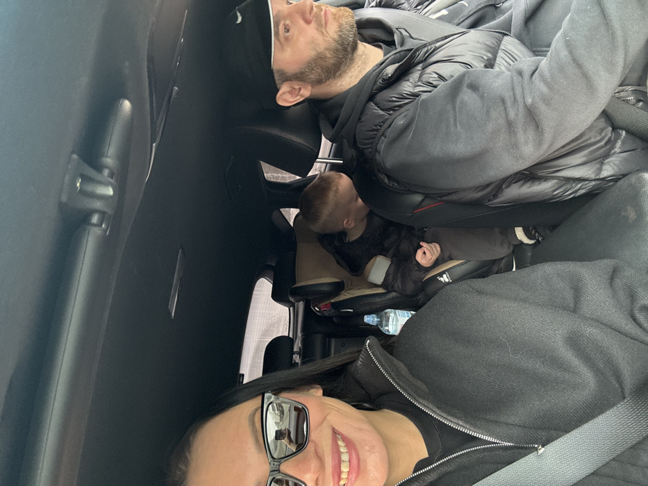 Within a vehicle in Kabanov Mys, Russia, three individuals are present. A female is positioned in the front-left seat, wearing black sunglasses that reflect a road scene, a dark zippered jacket, and a visible seatbelt; she is smiling at the camera. A clear plastic bottle with a blue cap rests to her right. In the front-right seat, a bearded male, wearing a black baseball cap and a dark puffy vest, is observed looking forward, indicating operation of the vehicle, with a seatbelt visible across his chest. A light-haired child is secured in a tan child safety seat in the rear-left, wearing a dark puffy coat, with its head bent forward. The vehicle's interior is uniformly dark, and external visibility through the windows is diffused.