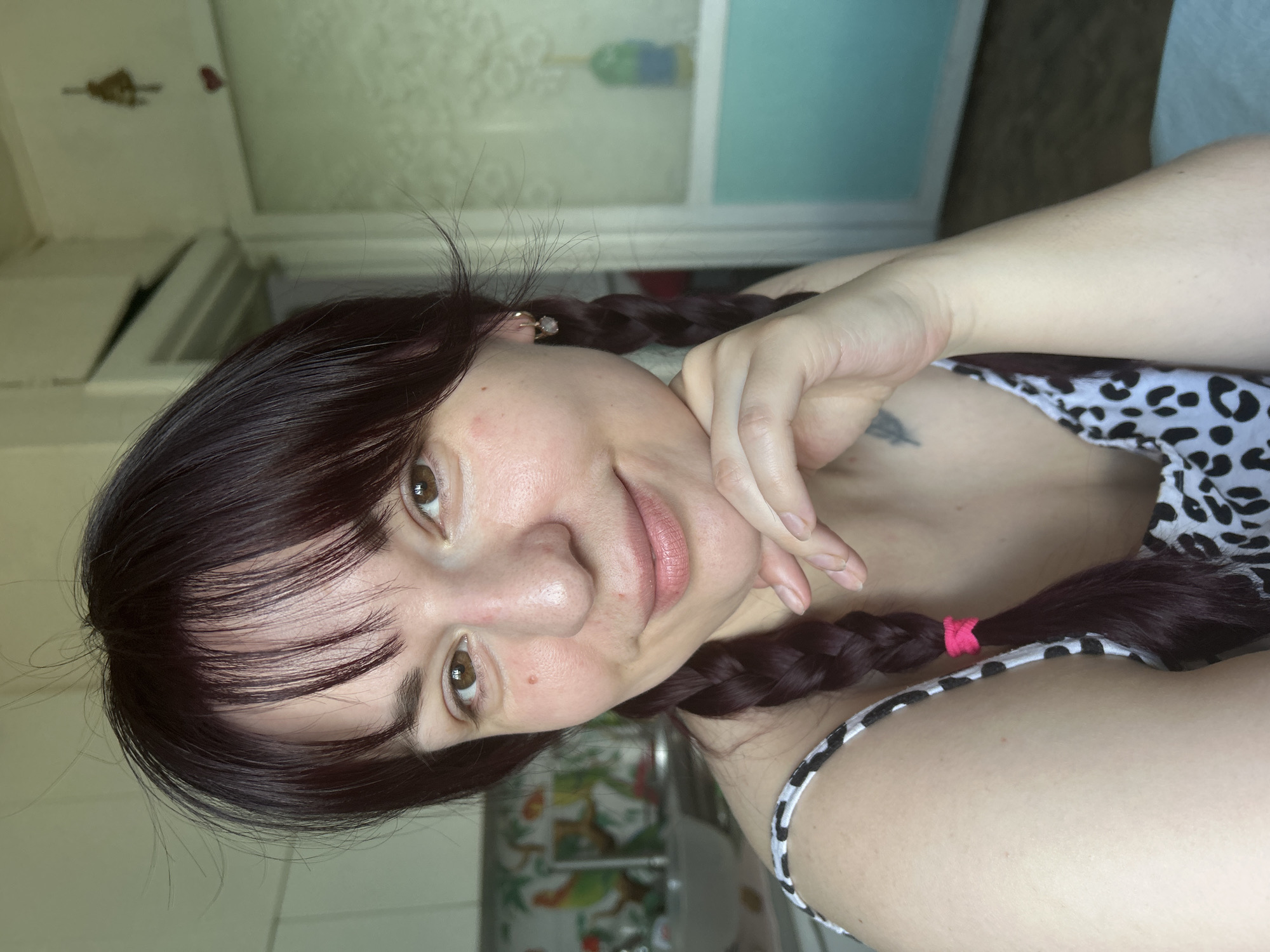 An adult female, appearing in her 20s or 30s, is captured in a front-facing, waist-up portrait in a residence in Фоки, Russia. She has dark, possibly burgundy-tinted hair styled with bangs and two braids, one of which is visible over her left shoulder and secured with a bright pink elastic band. Her skin exhibits a natural tone with minor facial redness visible on the nose and cheeks, and a small mole present on her right cheek. She has brown eyes and a subtle, light pink lip color, with a gentle smile directed towards the camera. Her right hand is positioned beneath her chin/jaw. She is wearing a sleeveless top featuring a black and white animal print. A small, light-colored charm hangs from a hoop earring in her left ear, and a faint tattoo resembling a feather or leaf is visible on her left upper arm. The background on the left reveals a kitchen area with white upper cabinets, a sink, a faucet, and a patterned backsplash displaying green and orange motifs. A white bowl is partially visible on the counter. The background on the right features a light-colored wall with a golden bell and a red heart decal. A doorway or opening leads to another room, where a blurry window or glass panel with a blue-green lower section and a bottle-like silhouette in green and blue hues is discernible. The floor in the distance appears dark.