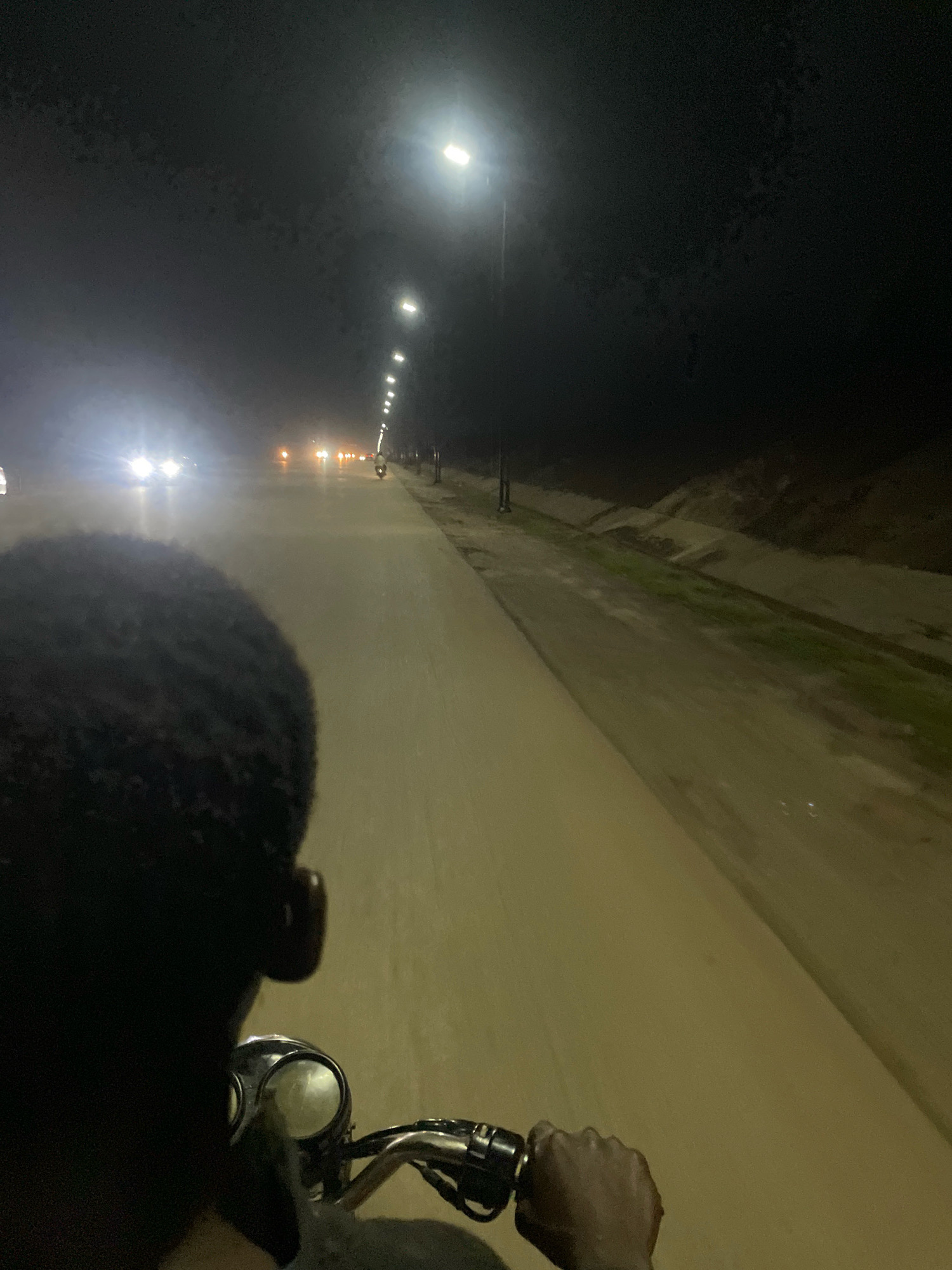 An over-the-shoulder, first-person perspective shot captures a person riding a motorcycle at night on an illuminated street, likely in Abuja, Nigeria. The rider's dark, close-cropped hair and the back of their head are visible in the foreground, obscuring their face. Their right hand is gripping the motorcycle's handlebar, which has a chrome accent and a black grip. The motorcycle's headlight, a circular lens with a bright, diffused light, is also visible.
The road ahead is paved and appears wet, reflecting the light from street lamps that line the right side of the road. These lamps are tall and modern, emitting bright white light. In the distance, several vehicles are visible, their headlights creating streaks of light on the wet road. The left side of the road features a grassy embankment with some evidence of erosion control or tiered landscaping. The overall environment is dark, with the primary light sources being the street lamps and vehicle headlights, suggesting a suburban or highway setting outside the most densely populated areas of Abuja. The atmosphere is consistent with nighttime driving in a region with moderate infrastructure.