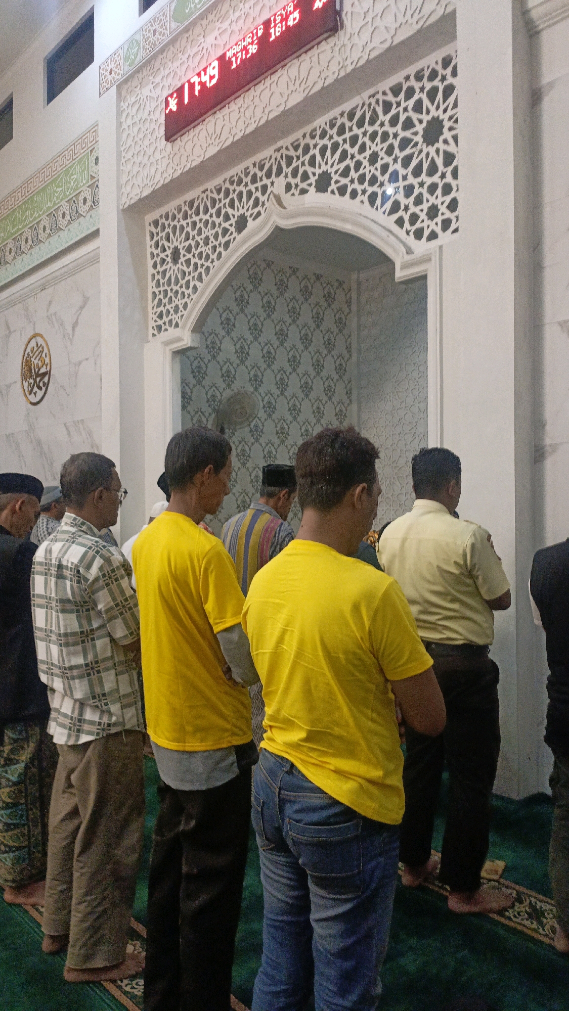 In the city of Ngaru Aru, Indonesia, a group of men are gathered in what appears to be a mosque. The men are standing in prayer, facing towards an ornate archway. Above the archway, a digital clock displays the time as 17:49, along with prayer times for Maghrib (17:36) and Isya (18:45). The mosque's interior features intricate geometric patterns on the walls and decorative tile work. Several men are wearing prayer caps, and one man in the foreground is wearing a yellow t-shirt and blue jeans. The lighting suggests it is late afternoon or early evening.