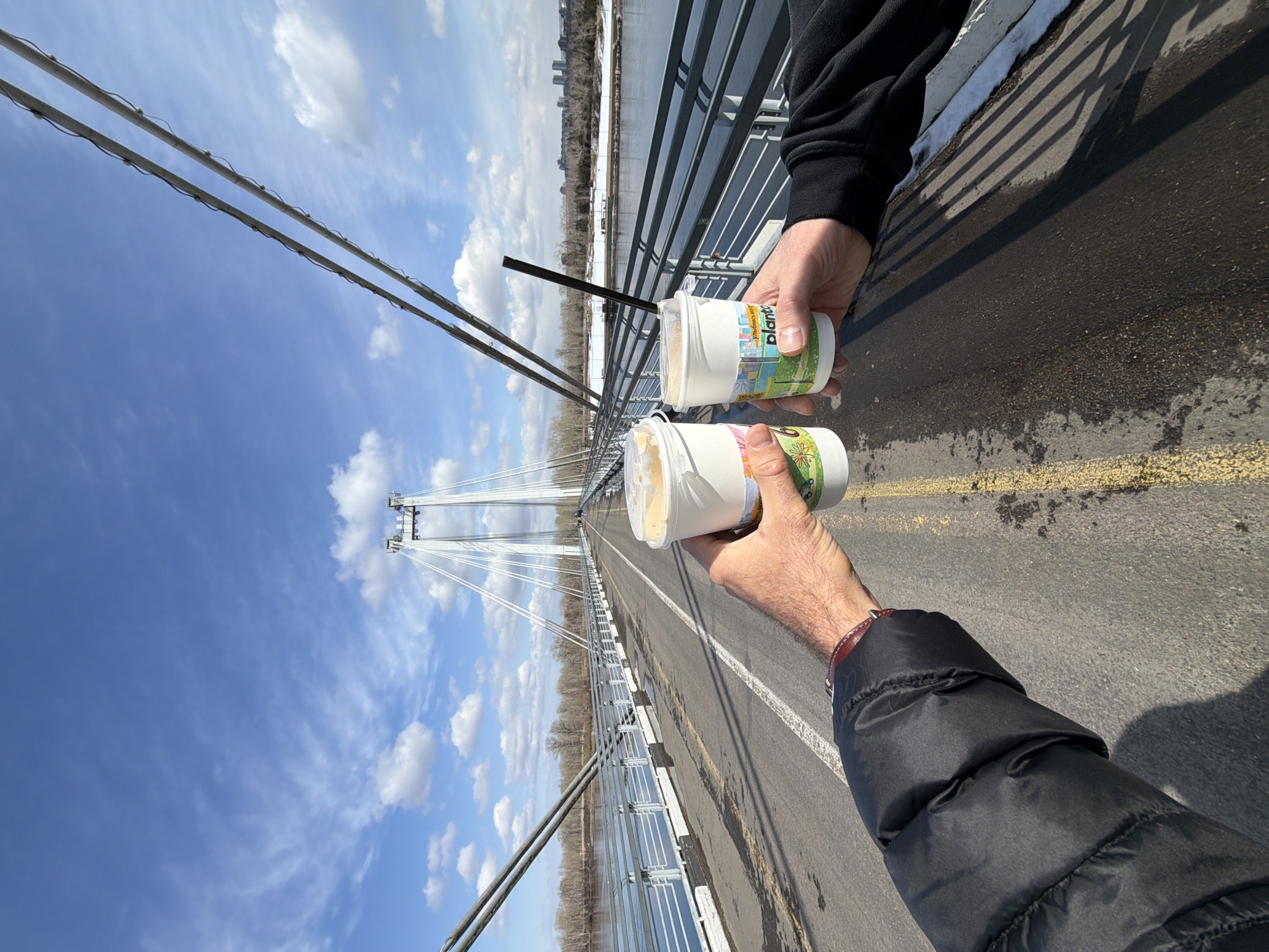 The image depicts two people toasting with cups on a bridge in Krasnoyarsk, Russia. The bridge is a cable-stayed bridge with a prominent central pylon. The sky is blue with scattered clouds. In the background is a body of water and the distant cityscape of Krasnoyarsk. The cups being held by the individuals appear to be cups of a beverage with straws, labeled "Planto". One individual is wearing a black long-sleeved shirt, while the other is wearing a black puffy coat with a leather-banded watch visible on their wrist. The surface of the bridge is visible, showing asphalt and painted lines.