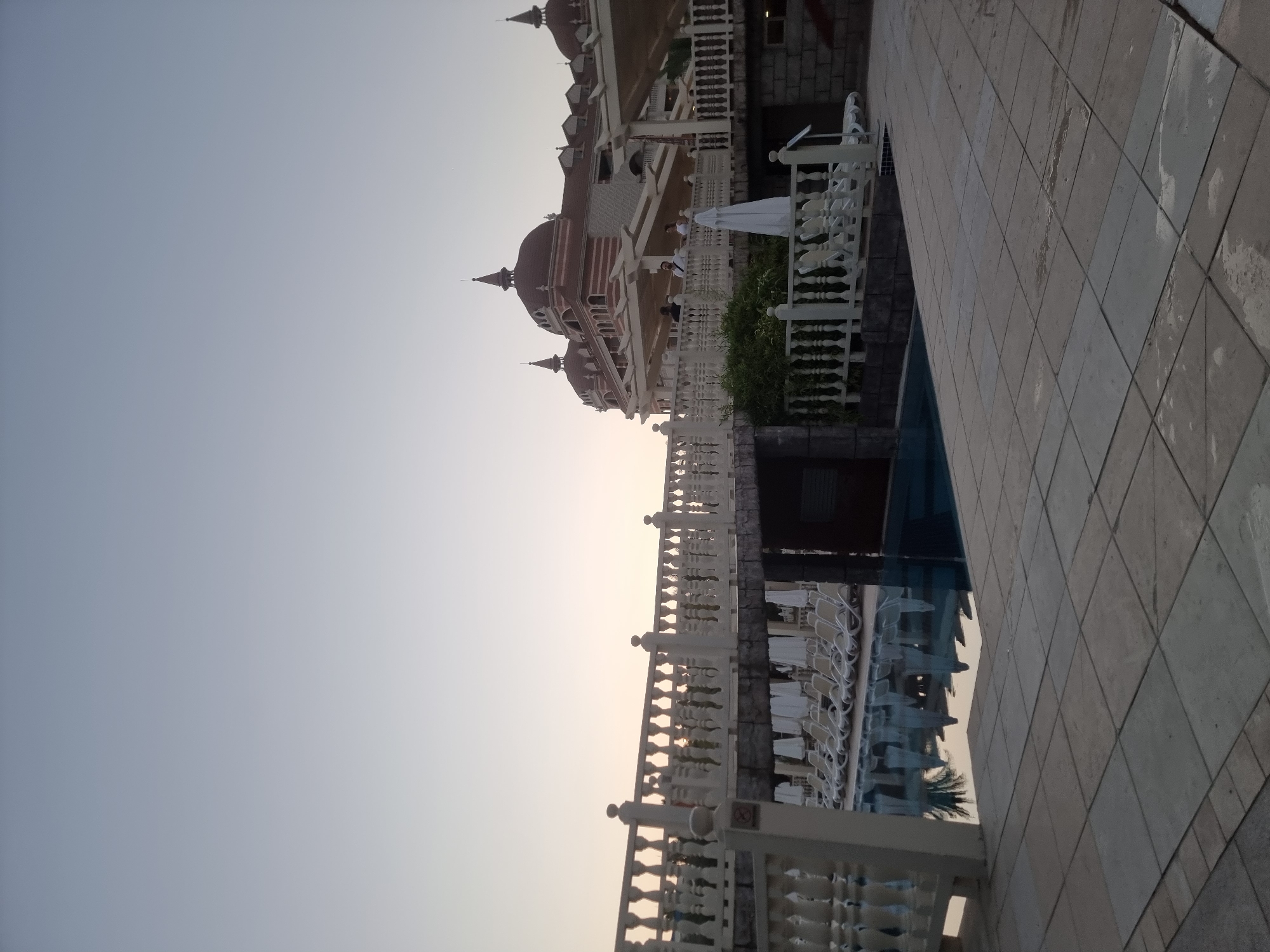 This media file captures a view of a resort or hotel with distinct architecture, featuring multiple domes and spires reminiscent of a palace or a traditional Middle Eastern style building. A white railing lines the foreground, separating a tiled walkway from a swimming pool area. The pool is furnished with numerous white lounge chairs, many of which are unoccupied. Green foliage is visible behind the railing, adding a touch of nature to the scene.
A few people are present, appearing to be guests or staff, dressed casually. One person is standing near the railing, and others are further back, closer to the building. The time of day appears to be late afternoon or early evening, as indicated by the soft, diffused sunlight and the warm hues in the sky. The weather seems calm and clear.
The tiled walkway is made of large, square tiles, some of which show signs of wear or water stains. The building itself is multi-story, with intricate architectural details. The presence of a swimming pool and lounge chairs suggests a recreational setting, likely a holiday resort. The location context of Manavgat, Turkey, hints at a warm climate and a tourist destination. There is no visible text in the media file.