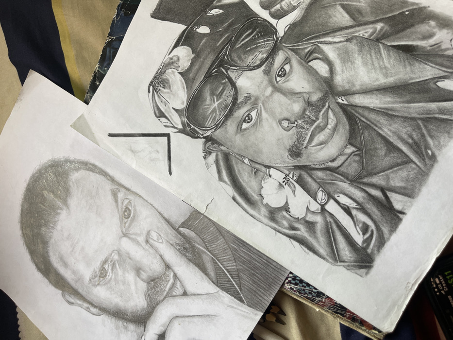 The image displays two realistic graphite pencil drawings of male portraits rendered on white paper, positioned in an overlapping arrangement. The location in Jos, Nigeria, cannot be determined from the visual evidence.
The upper-left drawing features a light-skinned male with short hair and a beard, looking directly forward. His left index finger is placed against his nose. He wears a textured garment, possibly a sweater. The paper shows slight creasing.
The lower-right drawing depicts a dark-skinned male with a mustache and a nose ring, also gazing directly at the viewer. He wears a dark bandana with a floral pattern and dark sunglasses positioned on his forehead. He appears to be wearing a jacket or collared shirt. This drawing's paper exhibits some wear and slight tearing along its lower and right edges.
The drawings rest on a blurred background of striped fabric, featuring dark blue and yellow horizontal bands. A portion of a dark-colored electronic device, possibly a mobile phone with faint white text on its screen, is partially visible in the bottom-left corner, beneath the drawings.