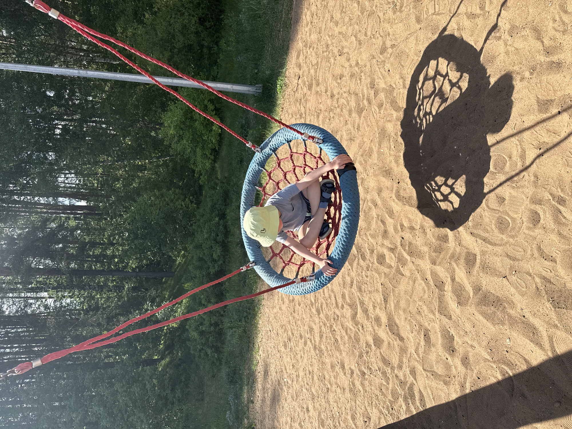 A child wearing a light green cap and a grey shirt sits in a blue-rimmed, red-netted swing in what appears to be a playground in Chaikovsky, Russia. The swing casts a shadow on the sand below. The playground is bordered by a green, wooded area, suggesting a park setting.