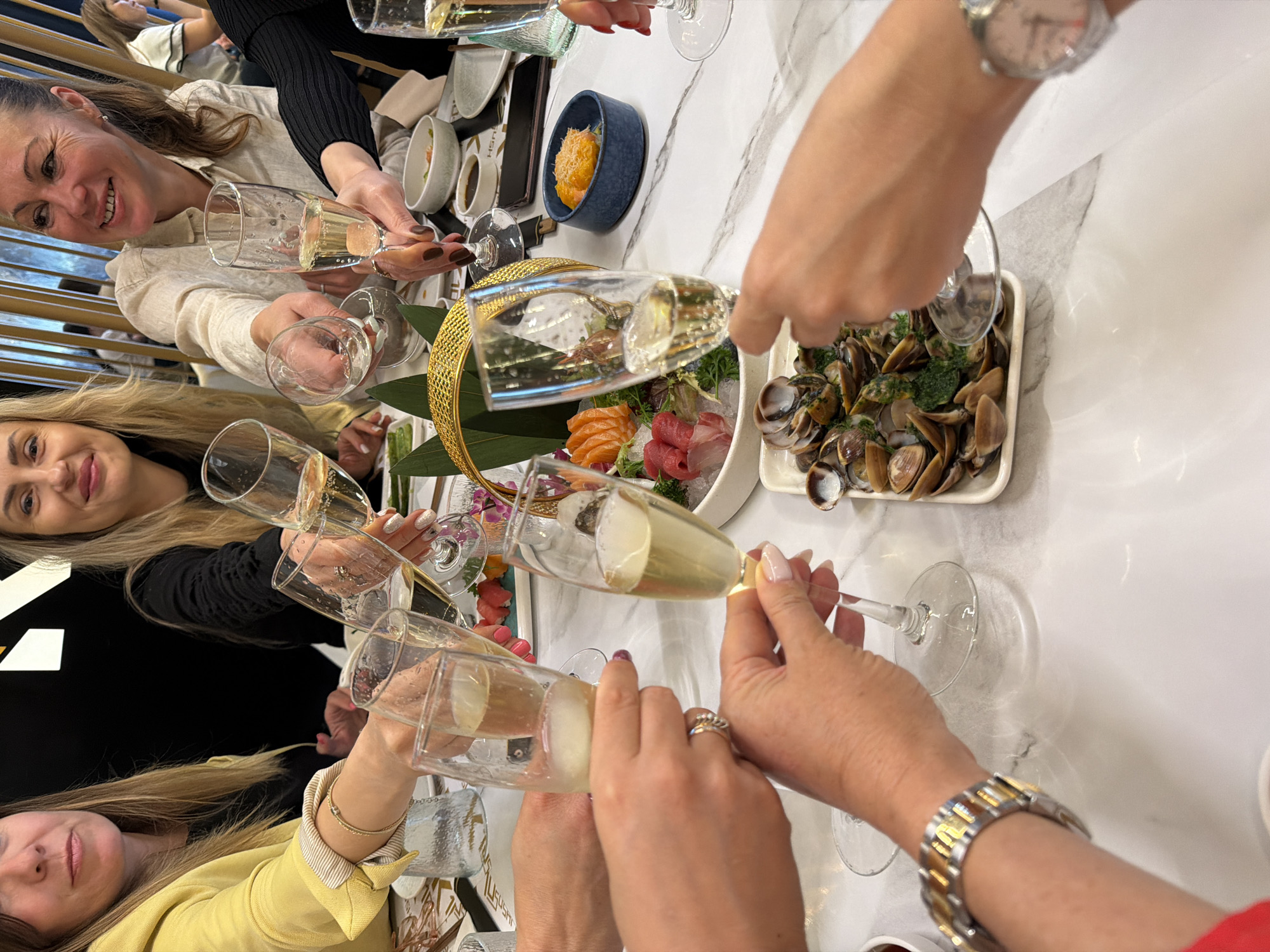 The image shows a group of six people toasting with champagne glasses around a table in an indoor setting. They are seated around a white, marbled tabletop on which are arranged a variety of dishes, including a sashimi platter, a bowl of orange roe, and a dish of clams. The individuals are dressed in various colors: yellow, white, and black. One person is wearing a watch with a white face and another a metal and gold bracelet watch. The table setting also includes additional plates and bowls in the background. The location appears to be a restaurant setting, though it is not possible to confirm its location in Tarragona, Spain.