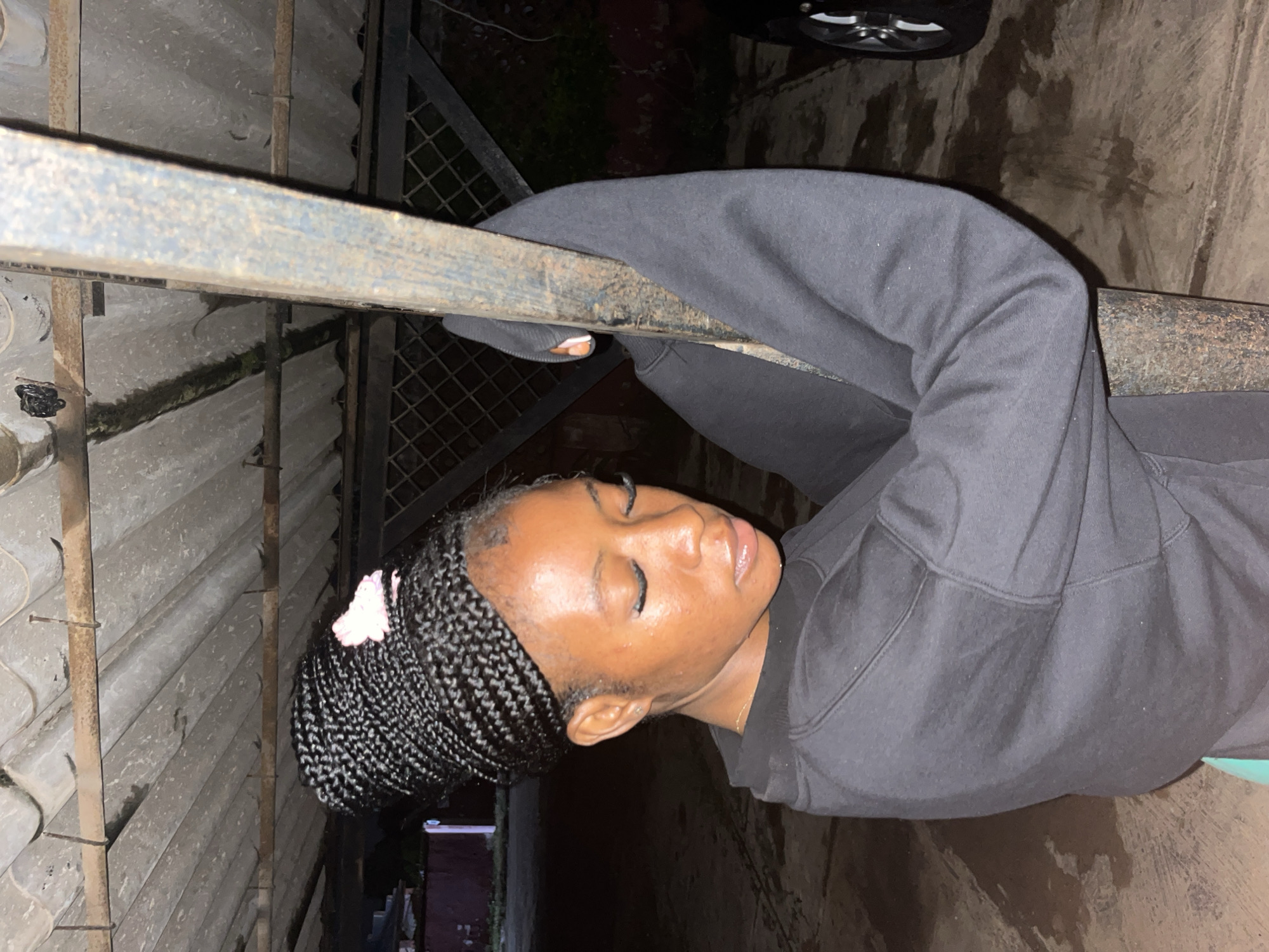A young Black woman with her hair in braided extensions piled high on her head and adorned with a small pink bow is leaning against a rusty metal pole. She is wearing a black hoodie and her eyes are closed. Her head is bowed, and she appears to be resting. The background suggests an outdoor location, possibly a parking area or under a covered structure, with a corrugated metal roof overhead and a chain-link fence visible. The ground is concrete and appears wet in places, indicating it might have recently rained. The overall scene has a slightly moody, informal feel. The image is taken at night, with artificial lighting illuminating the subject and her immediate surroundings. The location is identified as being in Jos, Nigeria.