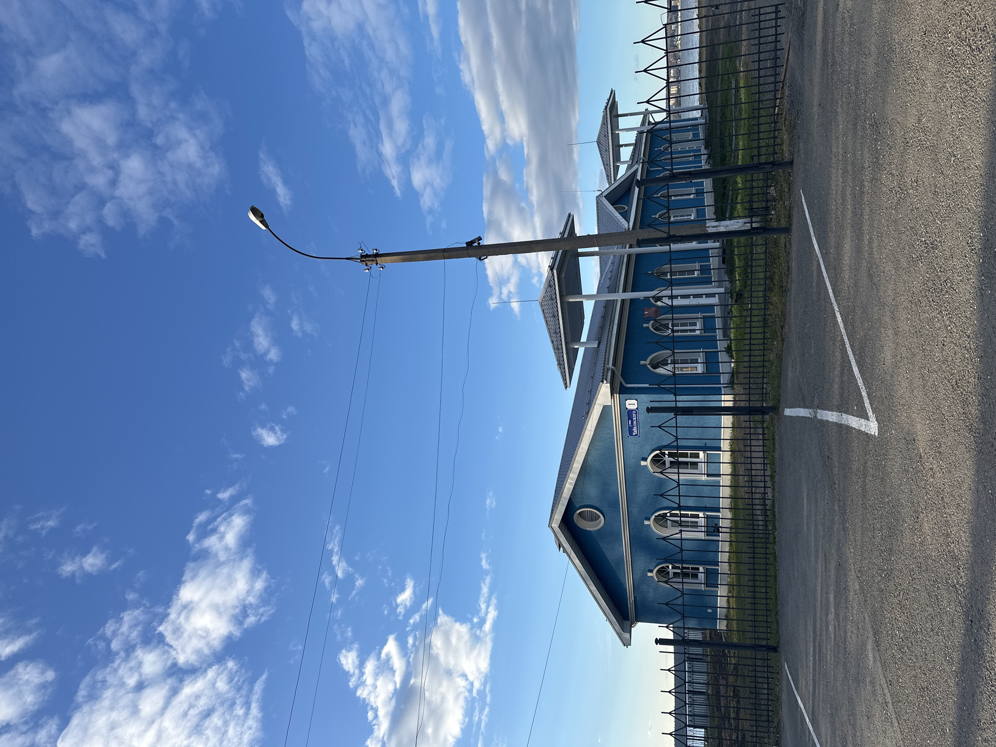 The image shows a blue building with white trim in Votkinsk, Russia, located adjacent to a paved parking area. The building has arched windows and a sign indicating "ул. Чайковского, 1" (Tchaikovsky Street, 1). A black metal fence surrounds the building. A utility pole with a streetlight is visible in the foreground, with power lines running across the sky. The sky is blue with scattered white clouds.