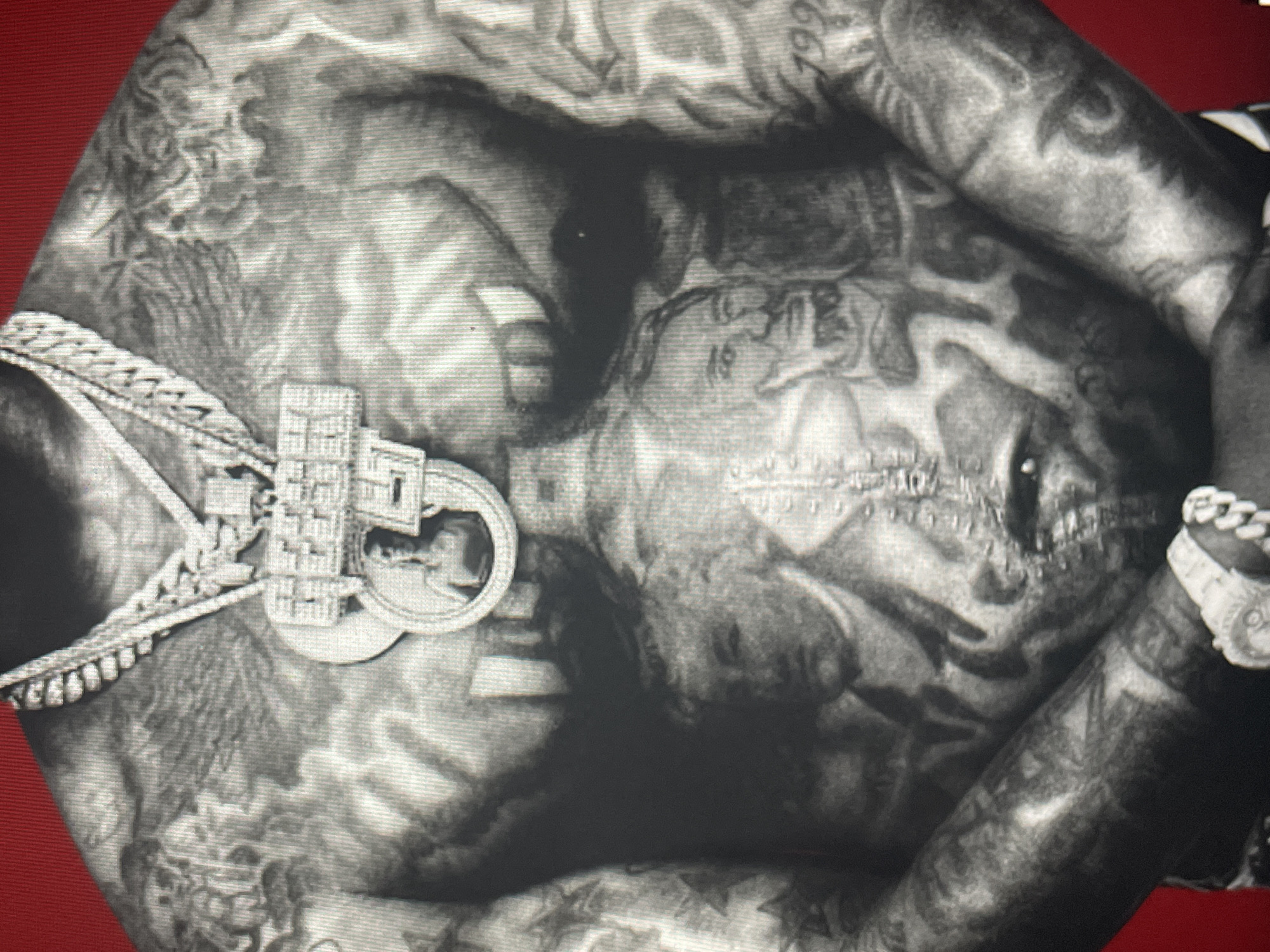 This close-up image presents a black and white portrait of an individual, likely male, captured from the upper torso to the top of the head. The person is heavily tattooed across their chest, neck, and face, with intricate designs that appear to include patterns, figures, and possibly words or numbers, though specific details are difficult to discern due to the image quality. On the person's right arm/shoulder, a tattoo displaying the number "199" is visible. A distinctive tattoo resembling stitches or scars runs across the mouth area.
The individual is adorned with prominent jewelry. A thick, diamond-encrusted chain is worn around the neck, featuring a large, ornate pendant. The pendant has a complex, geometric design, possibly depicting a building or symbolic structure, and includes a circular insert that shows the profile of a head, reminiscent of a coin or medallion. A matching, chunky, diamond-encrusted watch or bracelet is visible on the person's right wrist.
The image appears to be taken against a dark background, with vibrant red color peeking through on the top left and bottom right corners, suggesting a studio setting with a red backdrop or fabric. The person is posing, looking forward or slightly upwards, creating a confident or contemplative expression, though the heavy tattoos and grayscale rendering make precise emotional interpretation challenging. The overall visual style is striking and bold, emphasizing the tattoos and luxurious accessories. There are no visible cues within the image itself to suggest a specific location in Sofia, Bulgaria.