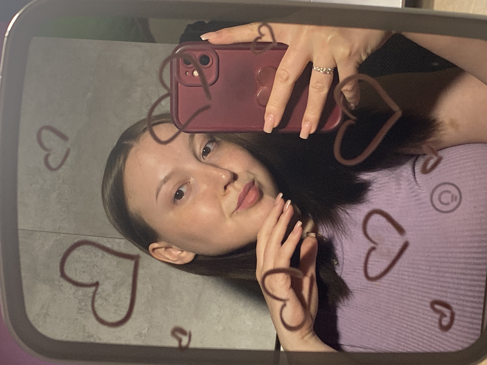 A young woman is taking a mirror selfie with a red phone. She is wearing a light purple top and a ring on her finger. There are heart shapes drawn on the mirror. The location is likely in an indoor setting, possibly a room in Chaikovsky, Russia.