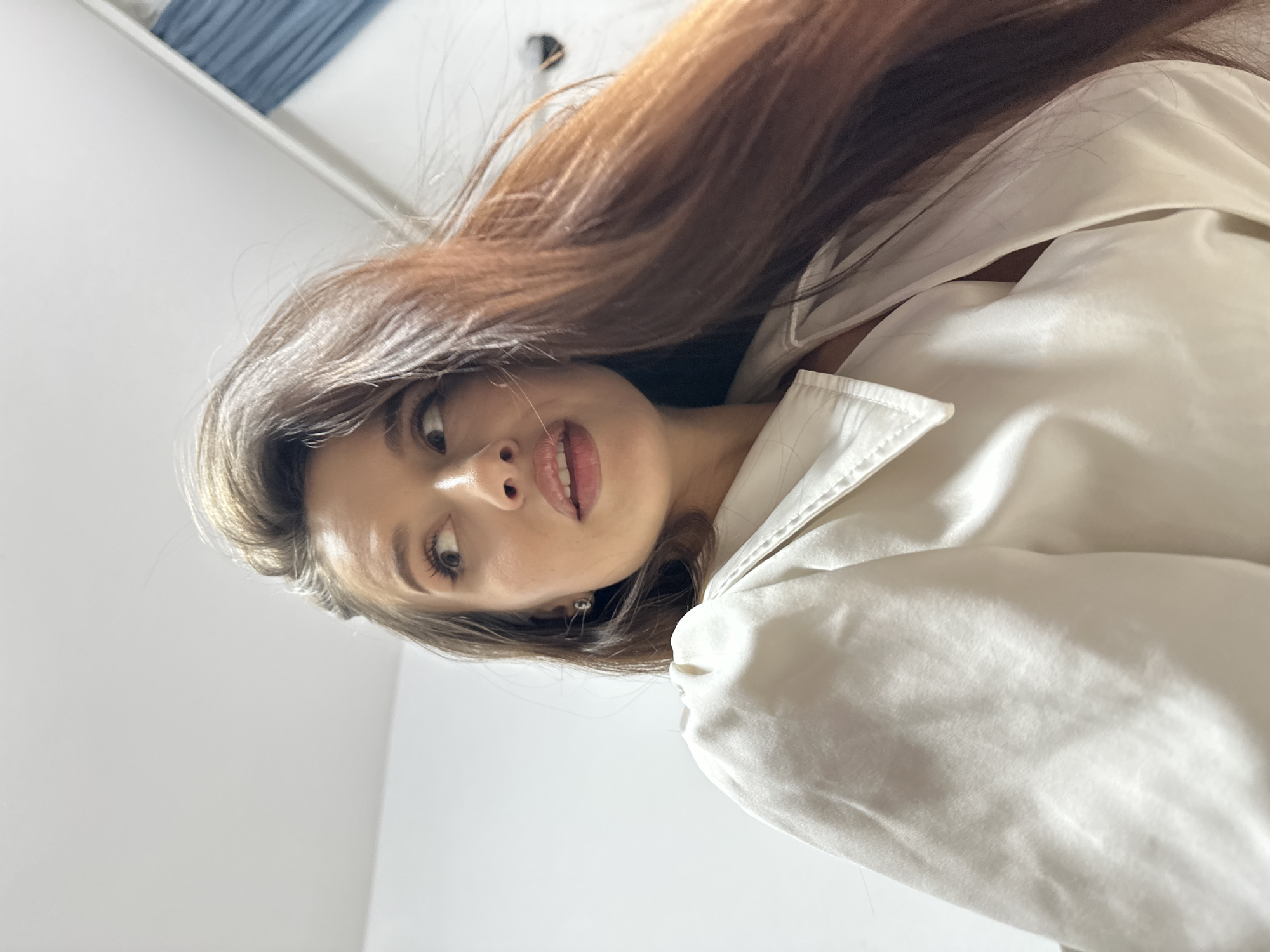 The image captures a close-up portrait of a young woman, rotated approximately 90 degrees counter-clockwise. She is looking directly down towards the camera, with a slightly parted smile revealing her teeth. Her long, reddish-brown hair flows across the upper and left portions of the frame.
The woman has fair skin, light-colored eyes, and well-defined eyebrows. She appears to be wearing subtle makeup. A small, shiny earring is visible in her right ear. She is dressed in a light-colored, possibly cream or white, collared garment, which appears to be a shirt or jacket, showing soft folds and a slight sheen in the fabric.
The background is predominantly white and minimalist, suggesting an indoor setting with bright, diffuse lighting. A hint of a blue object, possibly part of a bed frame or furniture, can be seen in the upper left corner of the rotated image. Her expression conveys a sense of direct engagement, curiosity, or gentle playfulness. No visible text is present in the image. The setting is indoors in Yekaterinburg, Russia.