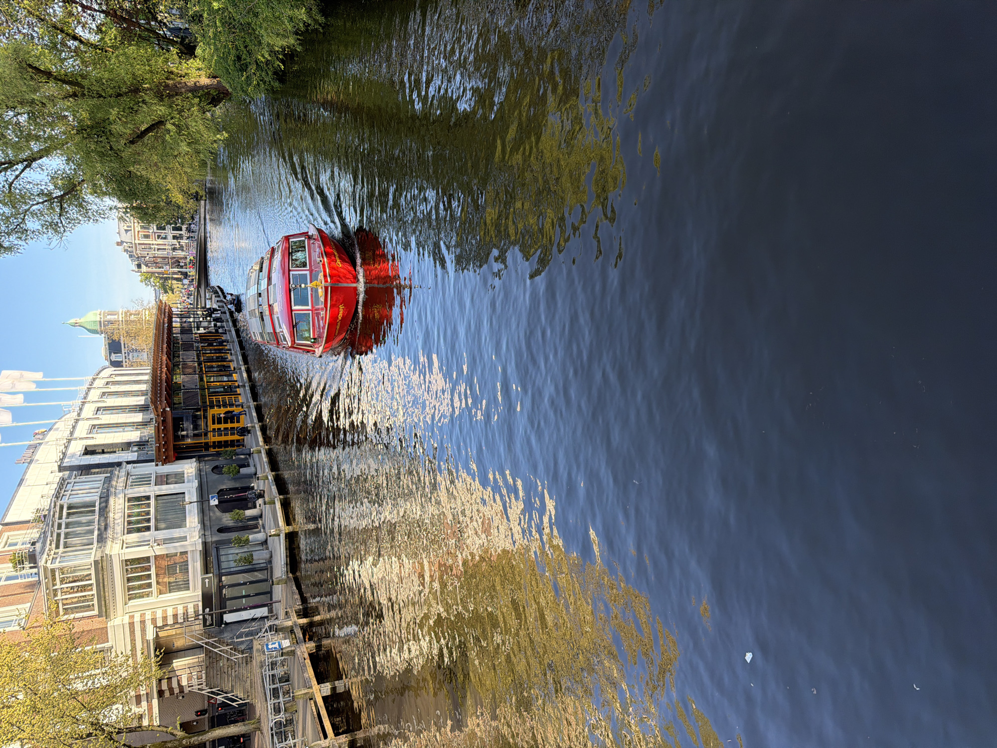 The image, oriented horizontally, depicts a red canal tour boat navigating a canal in Amsterdam on a clear, sunny day. The boat, positioned towards the center-left, is moving away from the viewer, leaving a distinct white wake on the dark water. Its red hull contrasts sharply with the canal's surface.
Along the left bank of the canal, a series of multi-story buildings, characteristic of Amsterdam architecture, are visible. These buildings feature varied facades, large windows, and some have street-level establishments or residences. Lush green trees line the canal, casting reflections onto the water. In the distance, a building with a distinctive green dome can be seen.
The canal water is a deep blue-grey, heavily reflecting the surrounding buildings, trees, and sky. The reflections are dynamic, particularly where the boat's movement creates ripples and shimmering patterns. Bright sunlight illuminates the scene, highlighting the water's surface and the boat's vibrant color.
Visible text on the side of the red boat clearly reads "LOVERS Canal Cruises," identifying it as a tourist vessel. On one of the buildings, a prominent "T" logo is visible, along with other indistinct signage. The time of day appears to be either morning or late afternoon, characterized by bright, clear lighting and subtle shadows. The overall scene captures a quintessential Amsterdam experience, with the canal serving as a vital thoroughfare for sightseeing.