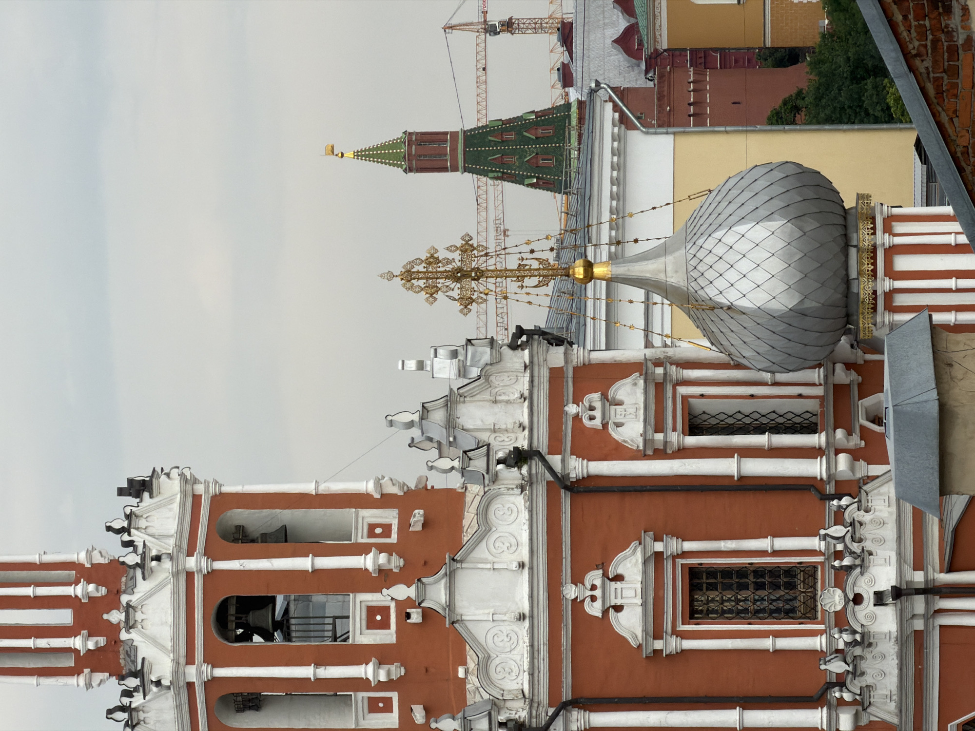 The image captures a view of the Moscow Kremlin with its distinct architecture. In the foreground, a red and white bell tower, characteristic of Russian Orthodox churches, dominates the left side of the frame. Its intricate white stucco details and arched openings housing bells are visible. To the right, a metallic, dome-shaped structure, possibly another part of the church complex, rises with decorative golden accents.
In the background, the green-tiled Spasskaya Tower of the Kremlin stands prominently, capped with a golden flag. Several construction cranes are visible, suggesting ongoing development or renovation within the city. The overall atmosphere is overcast, with a pale, diffused light. The scene is devoid of people, focusing solely on the architectural elements and the urban landscape of Moscow.