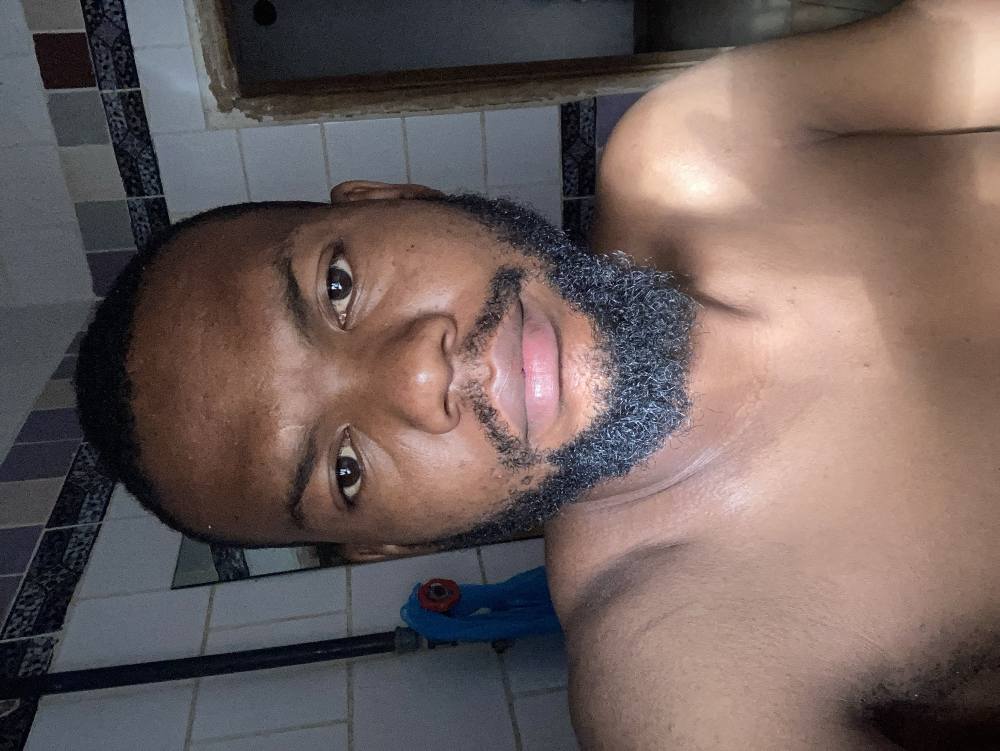 A close-up, rotated photograph shows the upper torso and face of a man. The man has brown skin, short black hair, and a graying beard. He faces the camera with a neutral expression. The background appears to be a bathroom, with white and patterned tiles visible on the walls. A metal pipe runs horizontally along the wall, and there's a partially visible blue object. It is not possible to confirm the location is in Jos, Nigeria from the image alone.