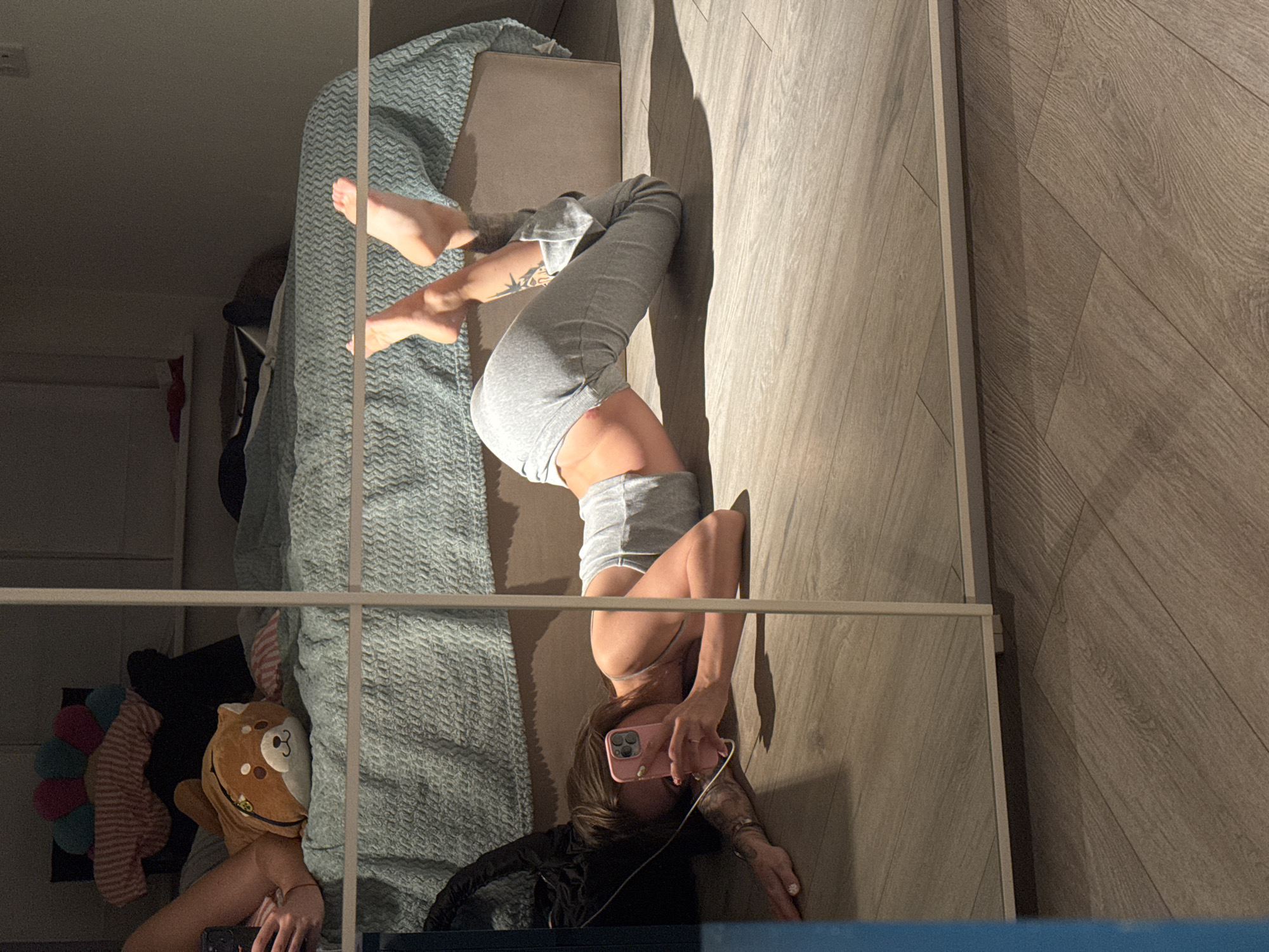 The image depicts a young woman with brown hair, lying on her stomach on a wooden floor, in what appears to be a bedroom. She is wearing a grey tank top and grey sweatpants, and is holding a pink smartphone, seemingly taking a selfie in a mirrored wardrobe. Her body is arched backward, with her feet pointing upwards. A stuffed animal resembling a Shiba Inu dog is visible on a bed behind her, which is covered with a blue textured blanket. The room is dimly lit, with sunlight casting shadows on the floor. The image is a reflection in a mirrored wardrobe, with its frame visible. The scene is located in Moscow, Russia.