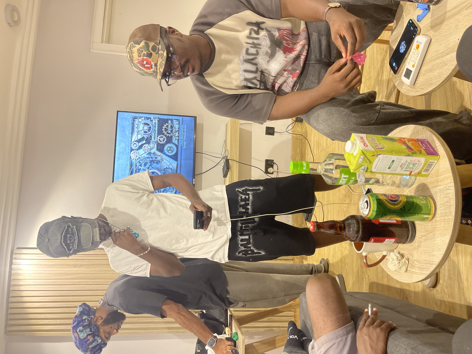 The image shows a group of people gathered around a coffee table in what appears to be an indoor setting, likely a living room or lounge area. The orientation of the image is rotated.
In the foreground, a person is seated, holding a cigarette. Another person standing next to them is wearing a dark t-shirt and a headscarf with a blue and purple pattern. Further back, two more individuals are visible. One is wearing a white t-shirt and black shorts with white lettering that reads "BALENCIAGA". They are holding a smartphone. The other person is wearing a camouflage baseball cap and a t-shirt with "ALACHIZA" printed on it, and they are holding what looks like a small pink object.
On the coffee table, there are several items including a carton of "Chivita Quotie" juice, a bottle of vodka labeled "FLIRT", a can of what seems to be beer, and a brown glass bottle. A white remote control and a smartphone are also on the table. A television screen in the background displays an image with gears and text that includes "Chris McClanahan" and "Google Featured photos", as well as a time stamp "17:15".
The lighting suggests it is daytime, possibly late afternoon. The overall atmosphere appears casual and relaxed, with the individuals engaged in conversation or activities around the table. The presence of drinks and electronic devices indicates a social gathering. The visible text on the clothing and products provides brand and content information.