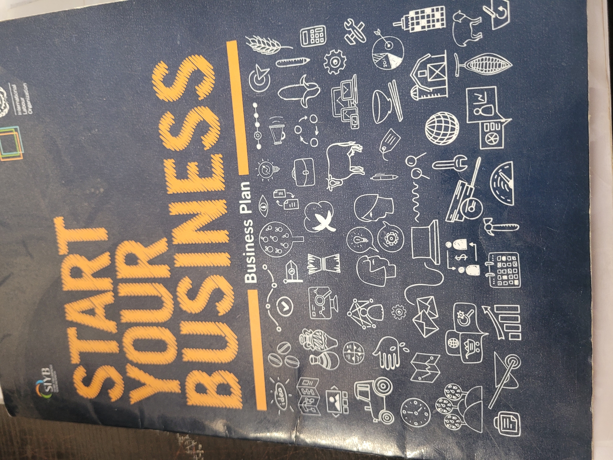 This media file displays the dark blue cover of a book or binder titled "START YOUR BUSINESS Business Plan." The main title "START YOUR BUSINESS" is rendered in large, textured yellow lettering, resembling embroidery or stitching. "Business Plan" is printed below it in plain yellow text.
In the top left corner, the "SIYB" logo is visible, accompanied by the vertically stacked text "International Labour Organization."
The majority of the cover, particularly the right side, is decorated with numerous small, white line-art icons that illustrate a wide array of business and economic themes. These icons include representations of:
- Agriculture: wheat, a cow, coffee beans, a cocoa pod, and cotton.
- Industry and Technology: gears, various tools, a factory, a truck, solar panels, computers, a laptop, a smartphone, a globe, and a light bulb containing the word "idea."
- Finance and Services: money bags, charts (one indicating "20%"), documents, a calculator with numbers like "8080" visible, a clock, a briefcase, figures of people, and communication bubbles.
The book cover appears slightly worn, suggesting use. No specific time of day or emotions are depicted, as the image focuses solely on the object itself. The item is located in Jos, Nigeria.
