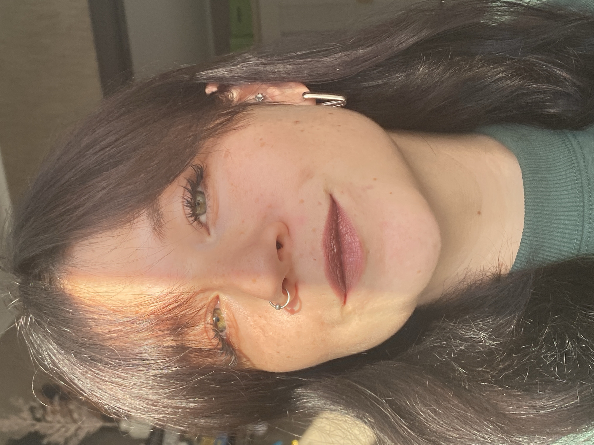 A close-up, head-and-shoulders shot features a young woman with dark brown hair and fair skin, adorned with freckles. She has green eyes and is wearing a nose ring and earrings. The lighting suggests natural sunlight, casting a warm glow across her face, with a portion of her forehead and right cheek illuminated by a strong light source. The background is blurred, showing hints of indoor decor. There is no identifiable location in the city of Chaikovsky, Russia, within this image.
