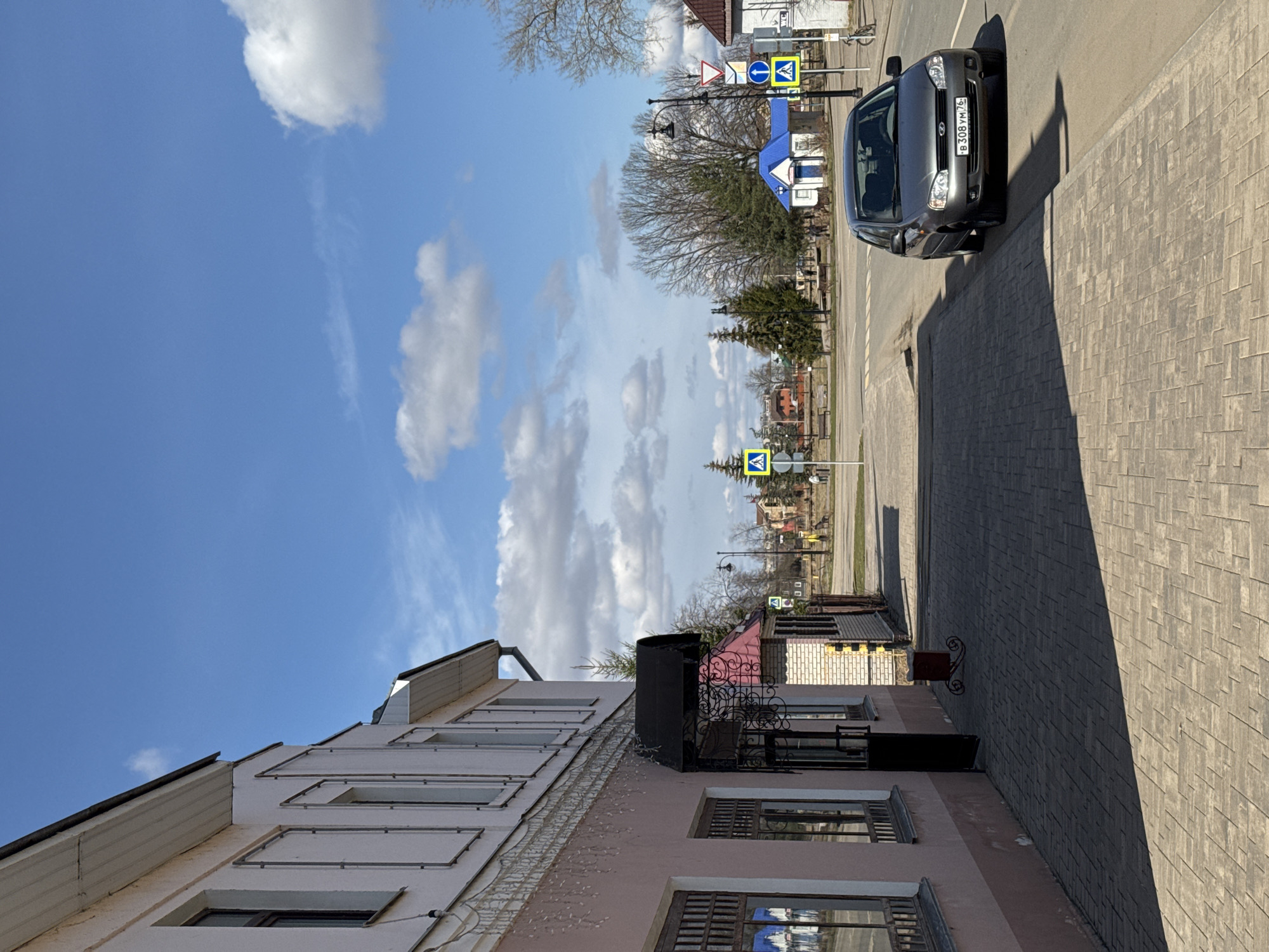 The image shows a street scene in Ново-Никольское, Russia. A grey car is parked on the right side of the road. Various street signs are visible along the road, including pedestrian crossing signs. A two-story building with multiple windows is on the left. The sky is blue with white clouds. The road is paved with grey blocks. The photograph appears to be taken at street level.