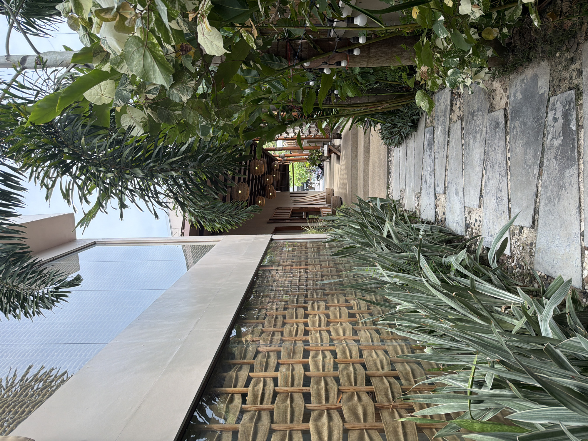 The image shows an outdoor pathway in Baan Bang Rak, Thailand. The pathway is paved with large, rectangular stone slabs. Lush greenery lines both sides of the path, including palm trees and various plants. A building with glass windows and a decorative woven screen is visible to the left. The pathway leads towards a covered area with seating and hanging light fixtures. In the distance, figures and vehicles are faintly visible. The overall setting suggests a resort or a tranquil outdoor dining area.
