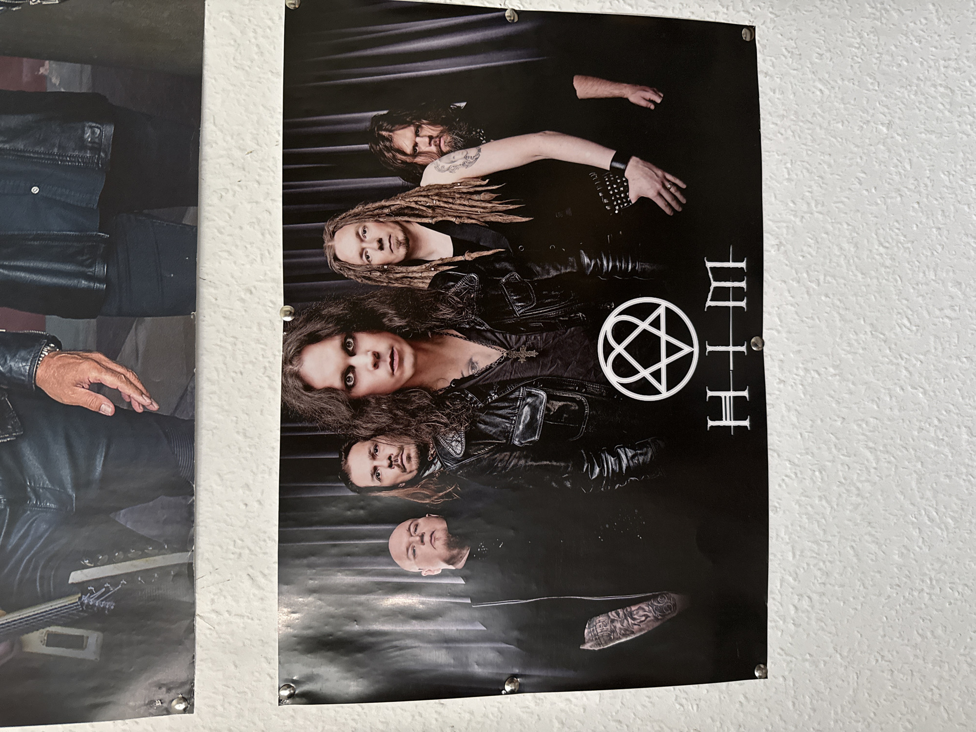 In the city of Bad Sachsa, Germany, two posters are visible, mounted on a textured white wall. The top poster, partially visible, shows a person's legs clad in black leather pants and boots, holding a guitar. The bottom poster displays a band posed against a dark curtain backdrop. Five men are present. The leftmost man has dark hair and a tattoo on his arm. Next to him stands a bald man wearing black. The central figure has long, dark hair and wears a leather jacket. To his right is a man with dreadlocks and a waistcoat. The rightmost man has long, dark hair and a beard. Below the band members, a white symbol of a heart with a double arrow through it is displayed, and the band's name, "HIM", is written underneath in stylized silver lettering. The posters are secured to the wall with metal fasteners.