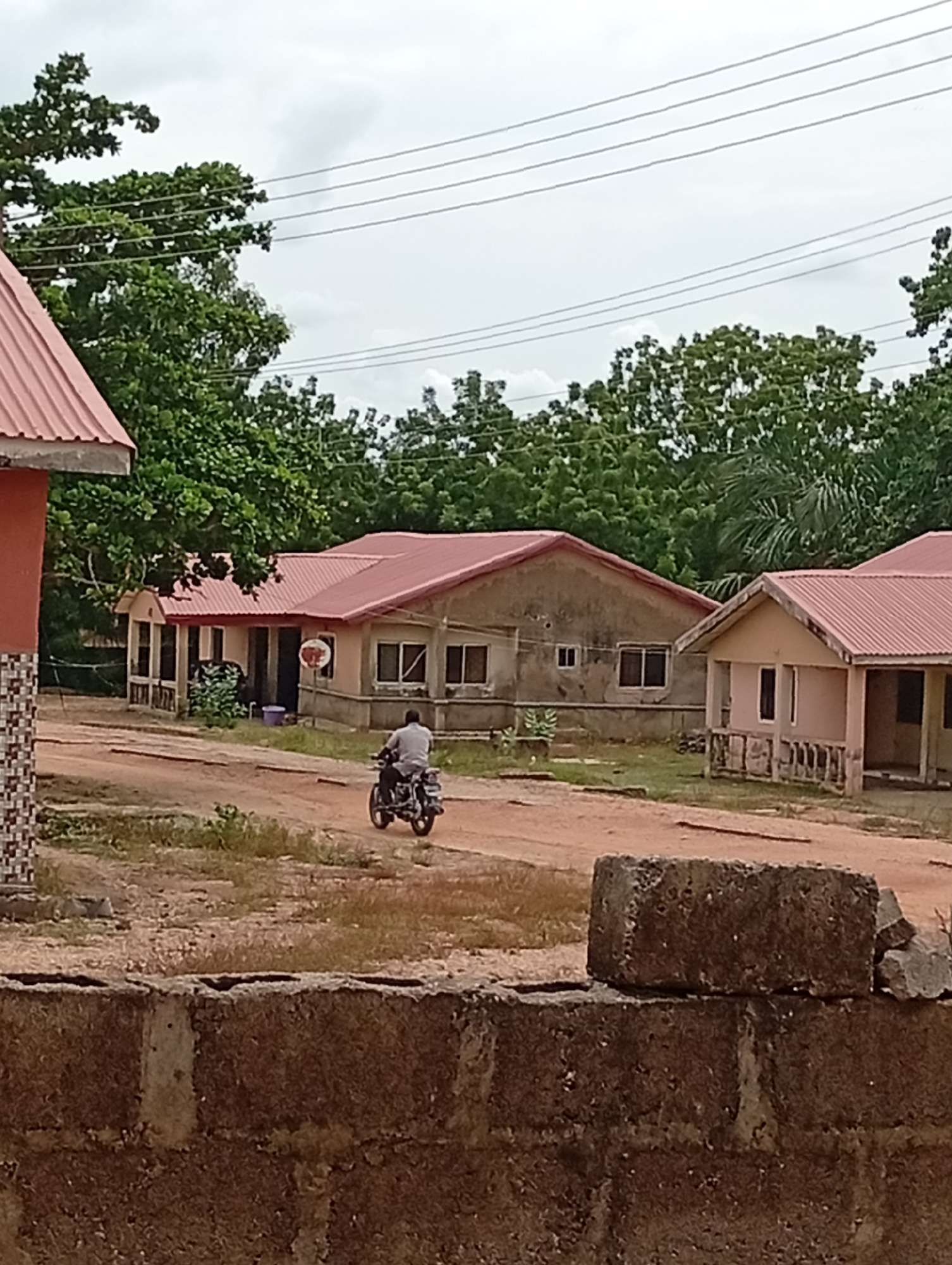 The image depicts a dusty, unpaved area in Adankolo, Nigeria. In the foreground is a roughly constructed cinder block wall. Behind it, a man is riding a motorcycle on a dirt road. Three single-story buildings are visible with reddish-brown roofs. Trees are visible in the background. Overhead, power lines stretch across a cloudy sky.