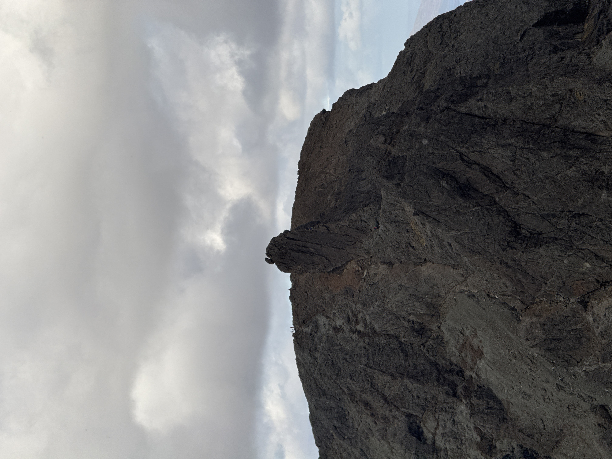 The image shows a rocky cliff against a cloudy sky. A few small figures, possibly people, are visible along the top edge of the cliff. The location is Unknown, Unknown.