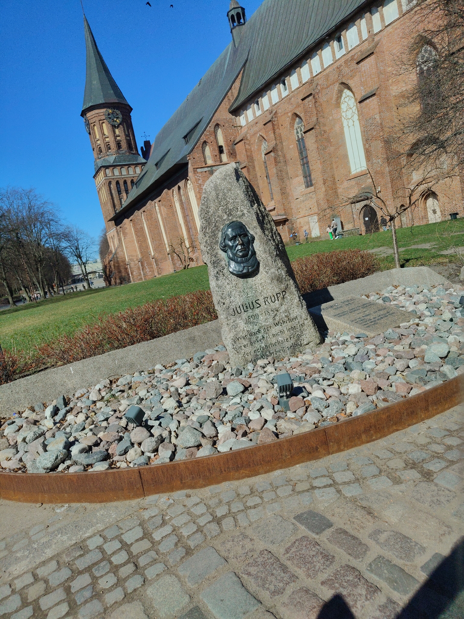 The image depicts a memorial stone dedicated to Julius Rupp, featuring a bust of the man and inscribed text. The memorial is situated in an area with grass, shrubs, and trees, adjacent to a brick building. The sky is blue with a few birds visible. The immediate surroundings of the memorial include a stone-filled area enclosed by a metal border and a cobblestone walkway. In the background, people are walking near the building.