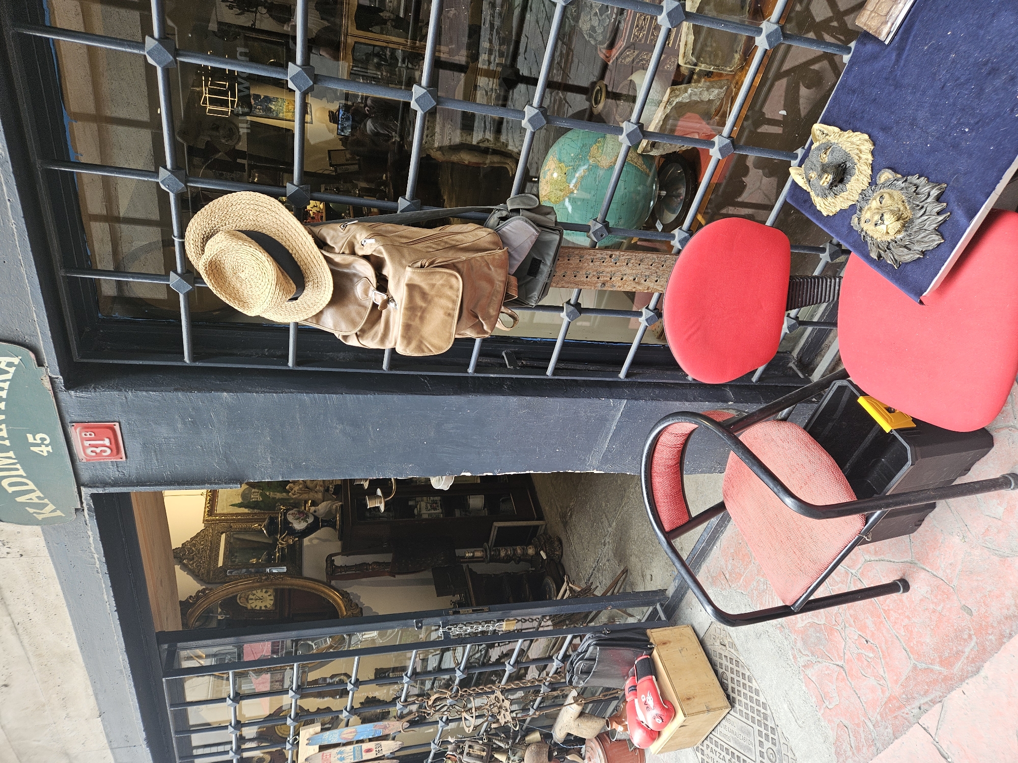 The image captures a scene at Kadim Antika 45, located at address 31B in Beyoğlu, Turkey. The view is from the outside of the antique shop, through a barred window display, and into the store interior.
In the foreground, various items are displayed on the window bars. These include a straw hat, a leather backpack, a globe, and a blue plaque decorated with lion figures. Two red cushioned chair seats are leaning against the window, and a black toolbox is placed nearby. A separate chair with a red cushion sits on the sidewalk, next to a wooden box with a pair of red boxing gloves.
Through the window, the shop interior is visible, filled with antique furniture, including clocks, mirrors, and other decorative items. The sidewalk appears to be made of stone.
