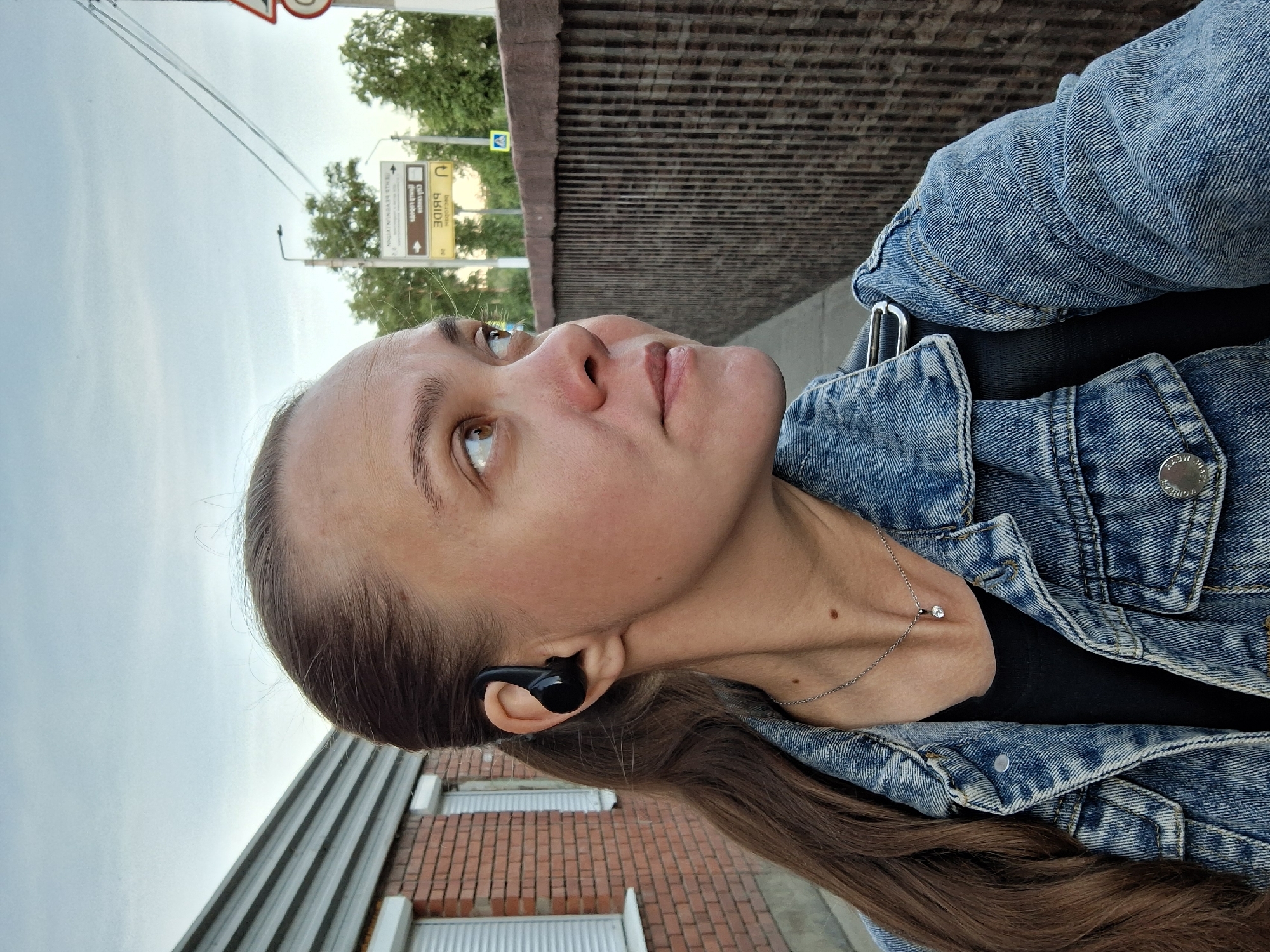 The image captures a candid, upright portrait of a young woman outdoors, looking upwards and slightly to the right. She has long, straight brown hair pulled back, and is wearing a blue denim jacket over a black top. A silver chain necklace with a small, clear pendant adorns her neck, and a black wireless earbud is visible in her left ear. Her expression is neutral and contemplative.
The setting appears to be an urban or semi-urban environment in Chaikovsky, Russia, likely a street or pedestrian area. In the background to her right, there's a tall, textured brick wall or bridge railing. Further back, trees are visible under a bright, overcast or hazy sky. To her left, part of a building with horizontal gray siding and brickwork can be seen.
Notable details include several road signs in the background. One brown rectangular sign has a white arrow pointing right, with partially mirrored and blurred Cyrillic text, and the English word "BRIDGE" clearly visible. Another yellow square sign features a U-turn symbol and also contains blurred Cyrillic text, with "ПРИДЕ" (Pride) partially decipherable. The time of day appears to be daytime, with soft, diffused lighting suggesting an overcast sky or perhaps late afternoon. The woman seems to be observing something above or ahead of her.