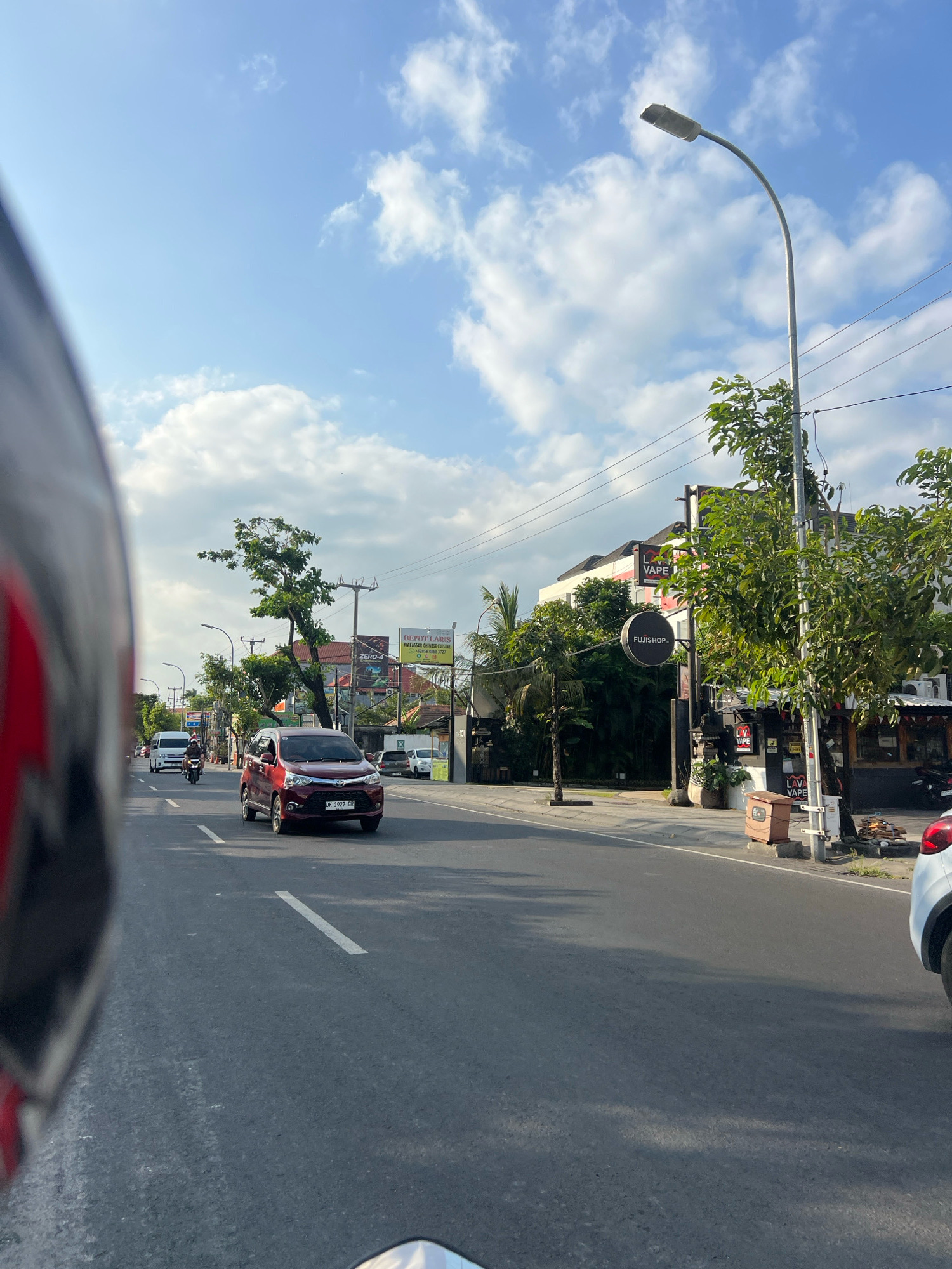 The image captures a street scene in Kuta, Indonesia, likely during the daytime given the bright sunlight and clear sky with scattered clouds. The perspective is from the viewpoint of someone on a motorcycle, as indicated by the prominent motorcycle helmet in the foreground on the left.
The road is paved and appears to be a multi-lane street. There is a clear white lane marking dividing the traffic. Several vehicles are visible on the road. A red car is in the right lane, moving away from the camera, and a white van is in the left lane, also moving away. A motorcycle with two riders is also in the left lane.
Along the right side of the road, there are buildings and shops. One prominent building has signs for "FUJISHOP" and "LAVA VAPE". Another sign above it reads "DEPOT LARIS MALAKASAN CHINESE CUISINE". Green trees and vegetation are present along the sidewalk and in front of the buildings, contributing to a tropical feel. A street light is visible on the right side of the road.
The overall impression is that of a typical urban street in a commercial area of Kuta, Indonesia, with ongoing traffic and businesses operating.