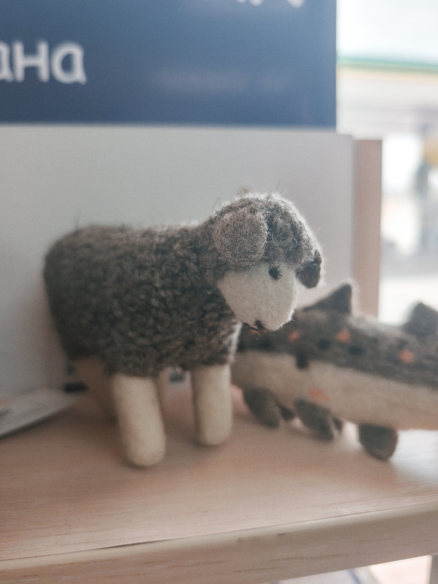 A felt sheep toy and another animal toy are displayed on a wooden shelf. Outside the window behind the shelf, a partial view of a gas station is visible. The sign partially displayed in the background says "HA" in Russian. The toys are indoors, likely a store or home, in Кашат, Kyrgyzstan.