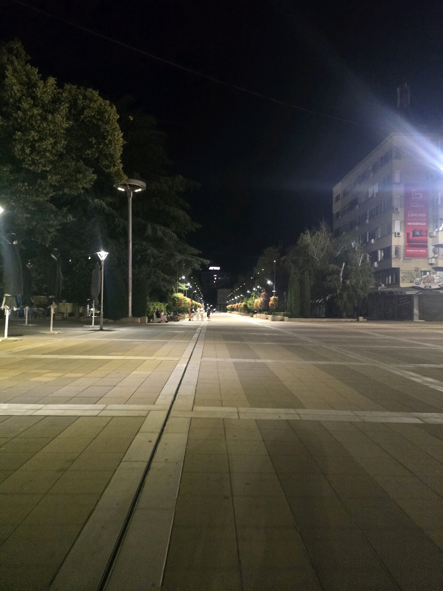 The image shows a pedestrian street at night in Yambol, Bulgaria. A wide, paved walkway with alternating light and dark tiles stretches into the distance. Streetlights illuminate the path, casting a warm glow. On the left side of the walkway, there are trees and outdoor seating areas, possibly for cafes or restaurants. On the right side, a multi-story building is visible, with a large advertisement on its facade. A few people are walking in the distance, and a motorcycle is parked near the building. The overall impression is of a quiet, urban evening scene.